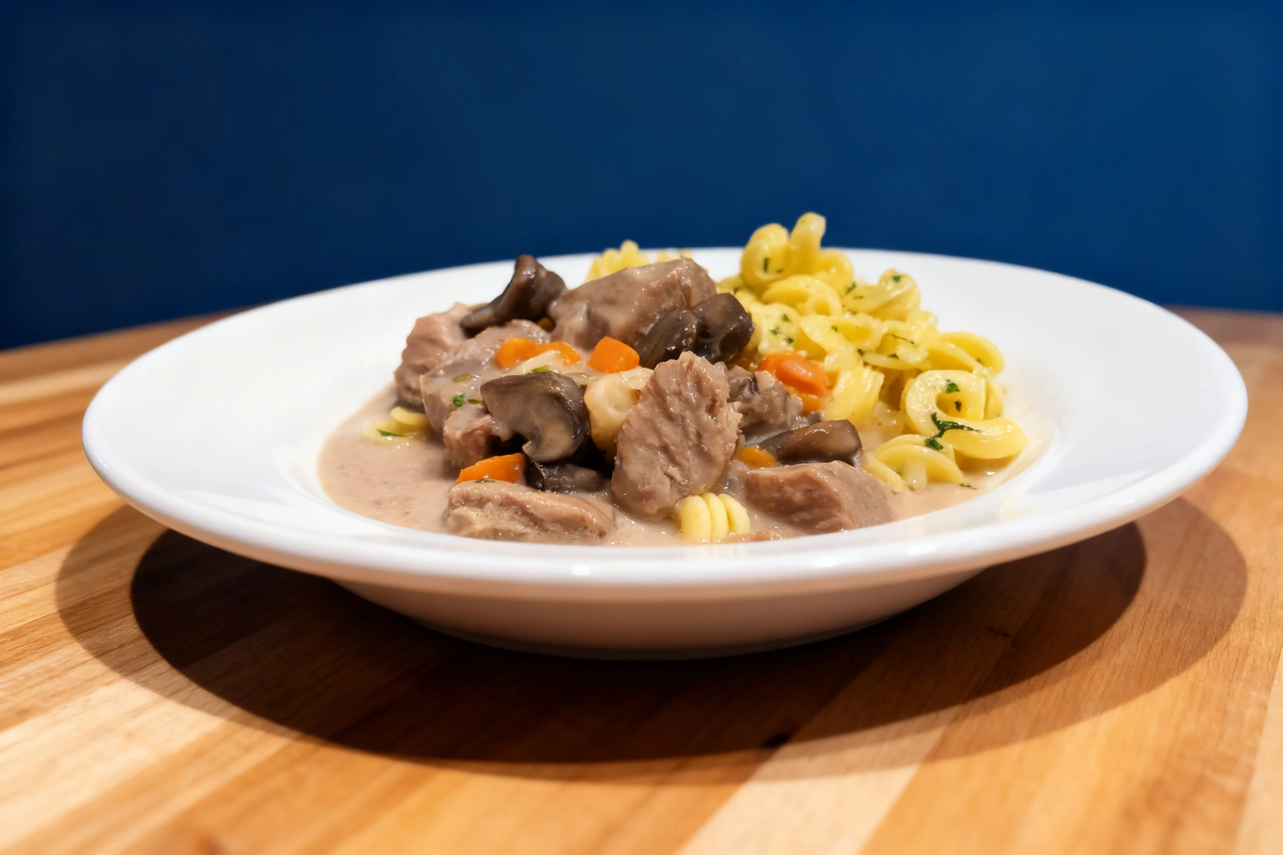 A meat stew featuring chunks of pork or beef with mushrooms and diced carrots in a creamy sauce, served with buttered rotini pasta garnished with herbs.