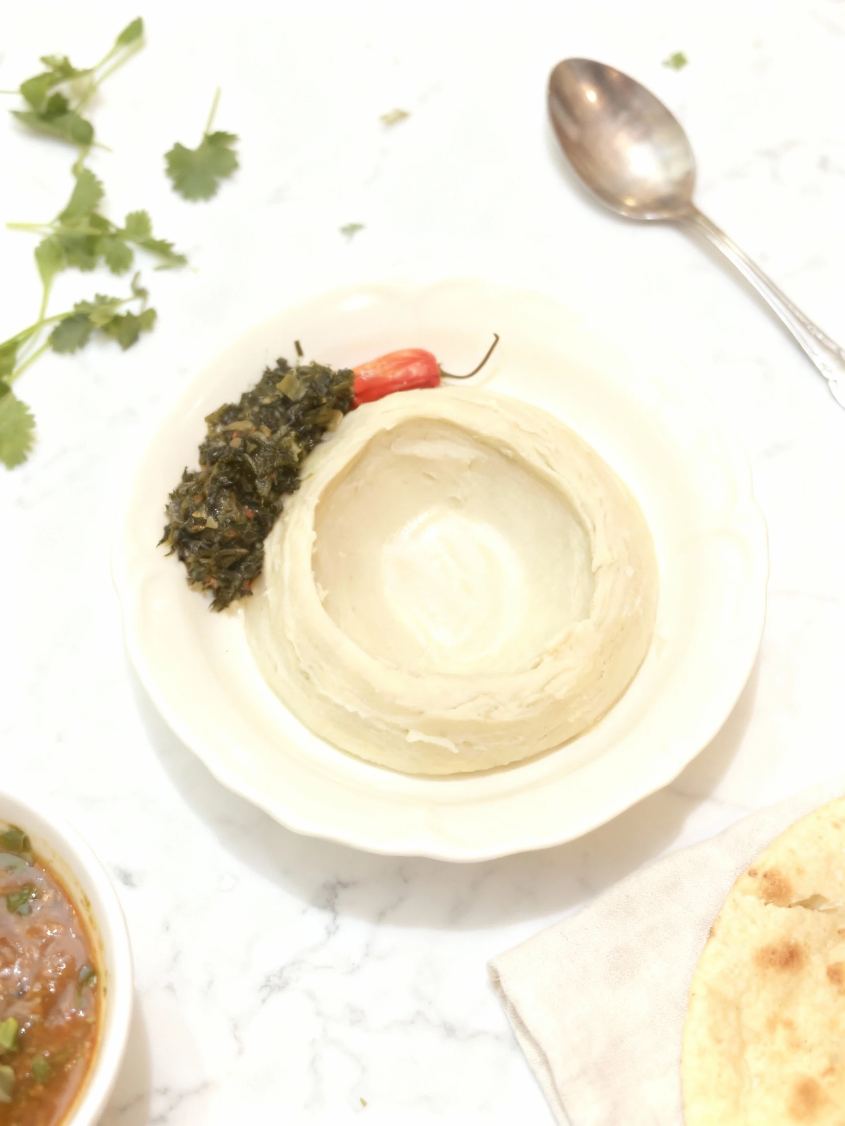 A molded serving of a pale, starchy staple (such as fufu or ugali) featuring a central depression, served alongside a pile of dark stewed leafy greens and garni