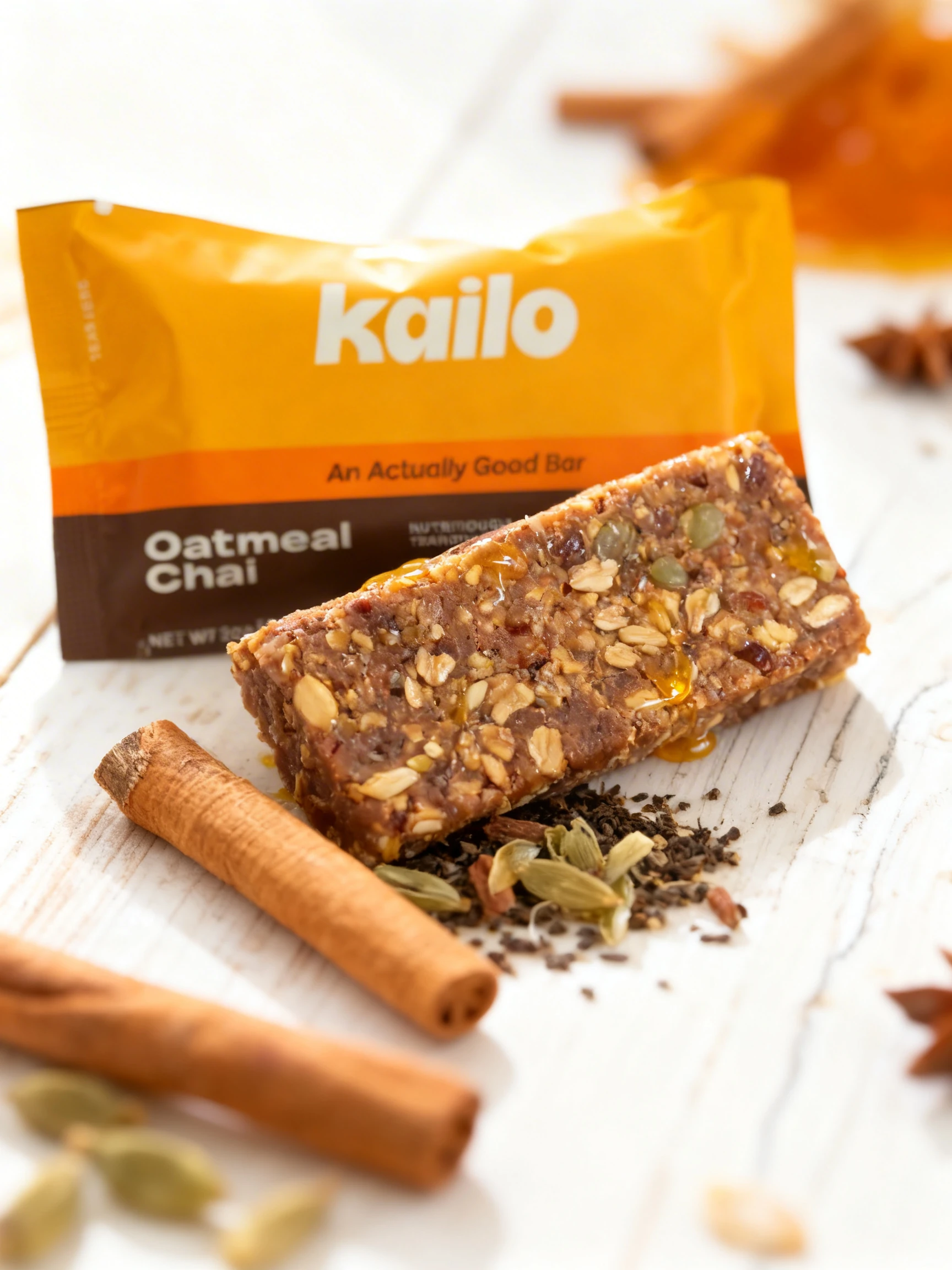 An Oatmeal Chai snack bar containing oats, seeds, and nuts, displayed next to its branded packaging with a cinnamon stick, cardamom pods, and loose tea leaves.