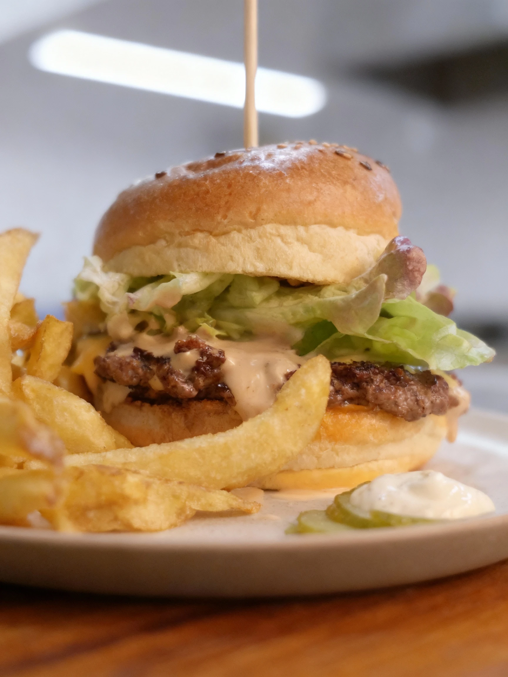 A sesame seed bun burger features a beef patty with melted cheese, fresh leaf lettuce, and a generous drizzle of creamy sauce, secured with a wooden skewer. It