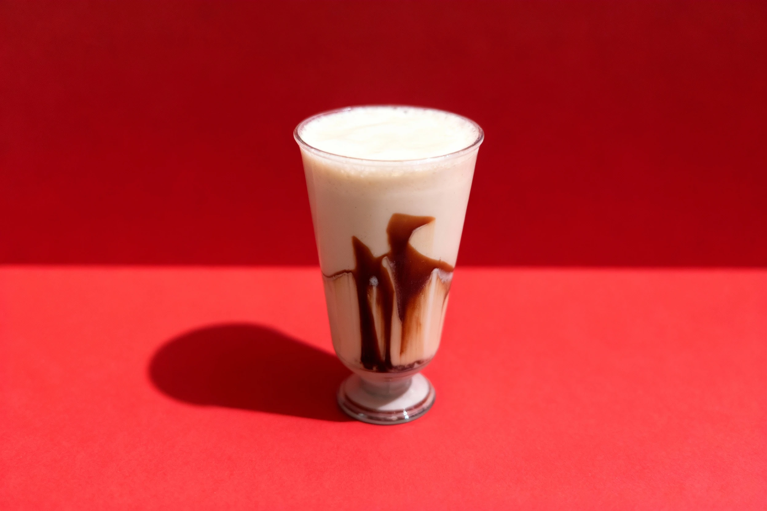 A creamy milkshake or beverage served in a clear plastic takeaway cup, featuring dark chocolate or caramel syrup heavily drizzled down the inside walls.