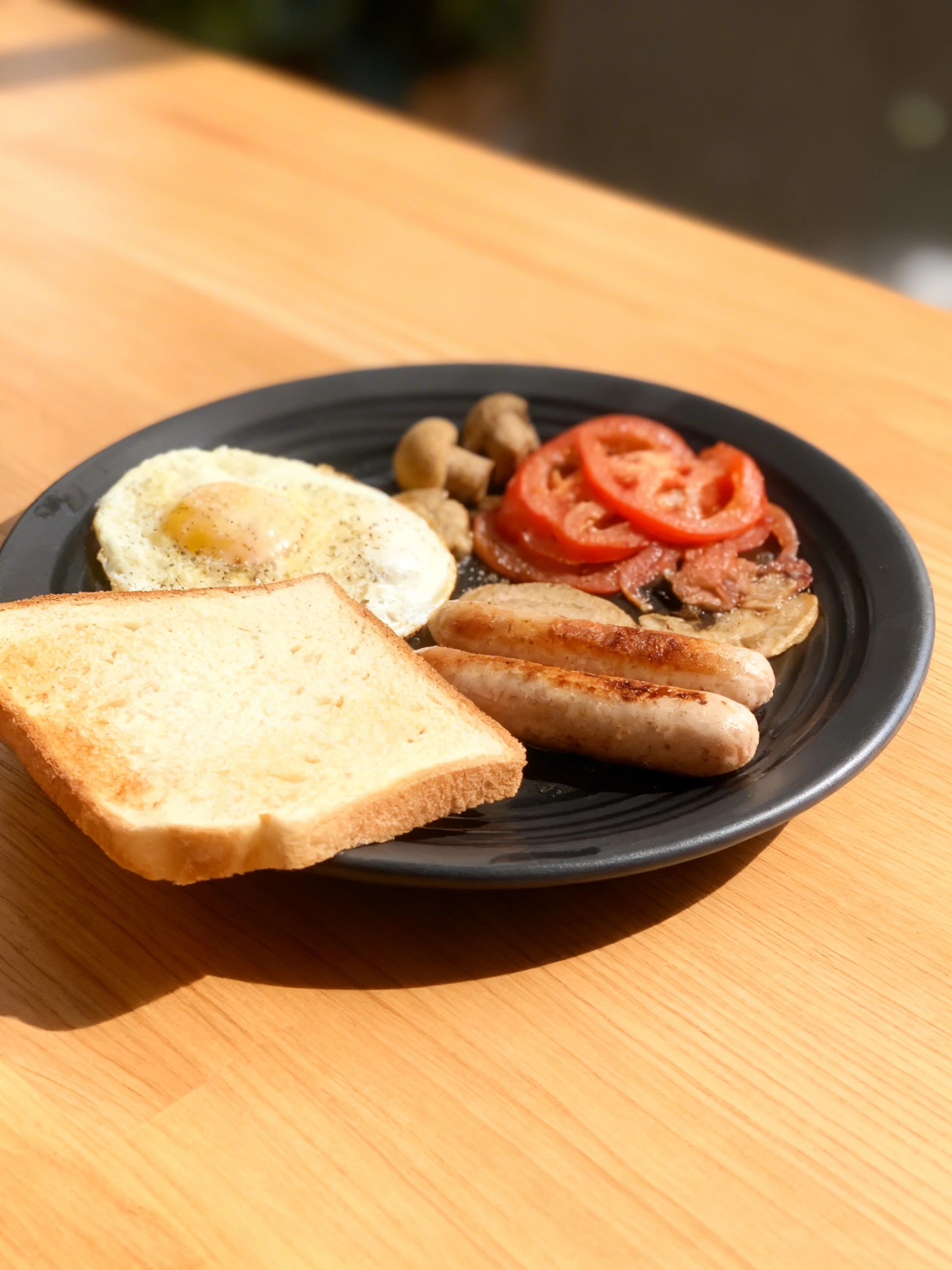 Breakfast platter featuring toasted white bread, a fried egg, and grilled sausages, accompanied by a small pan of whole mushrooms and sliced tomatoes.