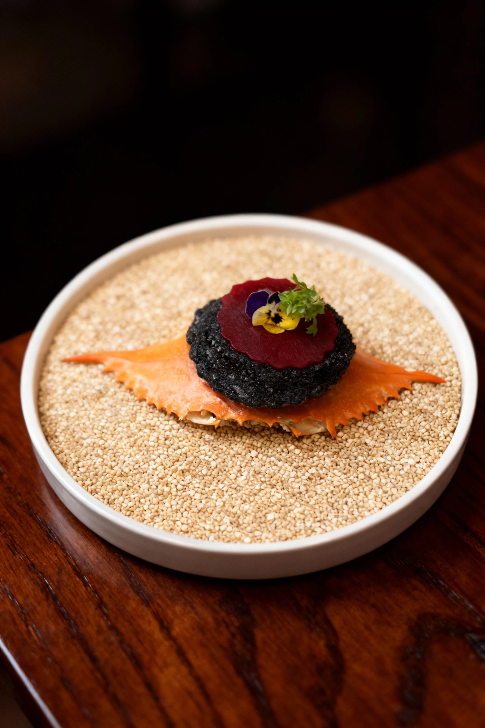 A black squid ink tartlet topped with a beet disc and edible flowers, served on top of a crab shell resting on a bed of grains.
