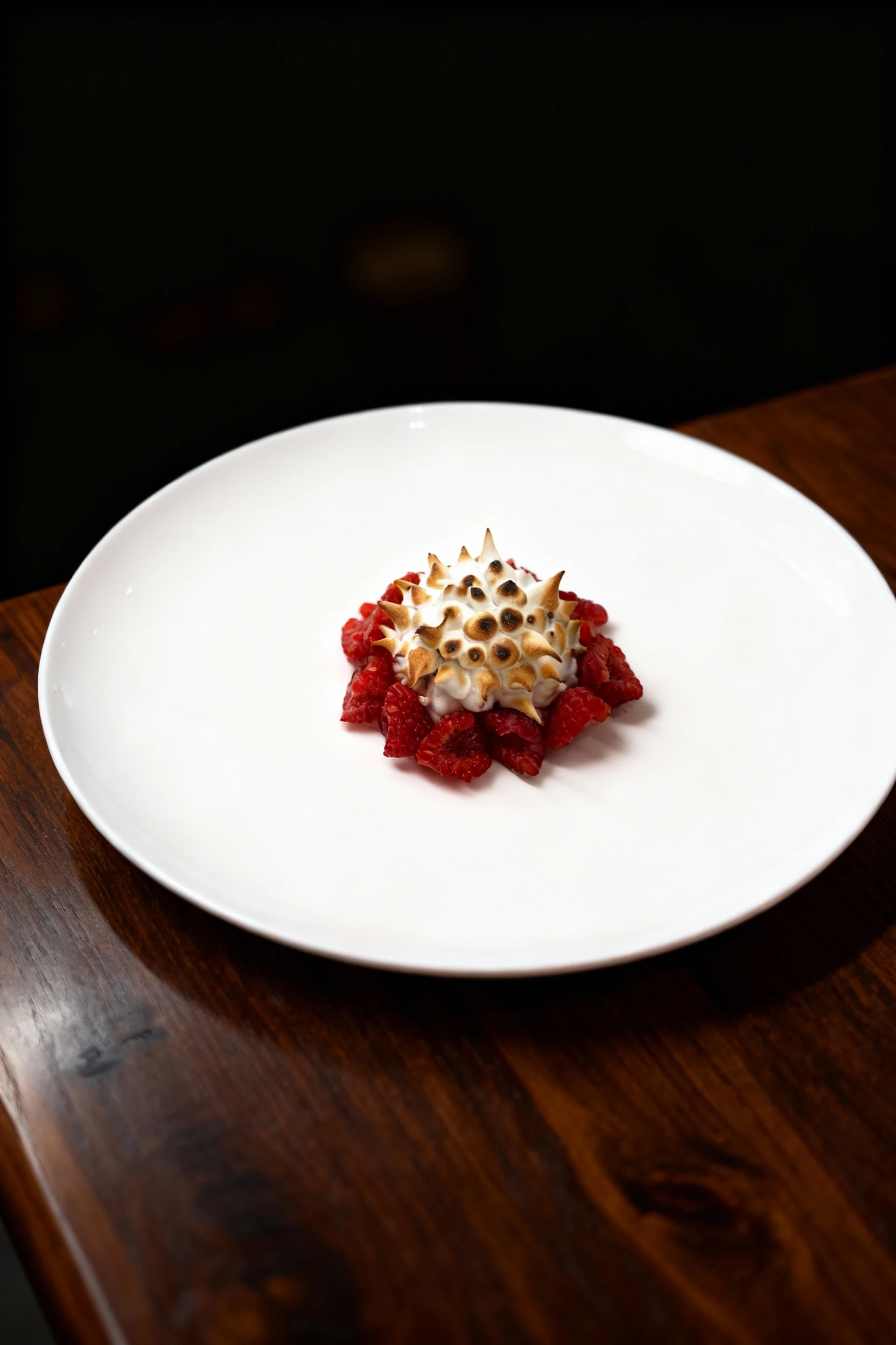 A plated dessert featuring a spiked mound of toasted meringue surrounded by a ring of fresh halved raspberries on a white plate.