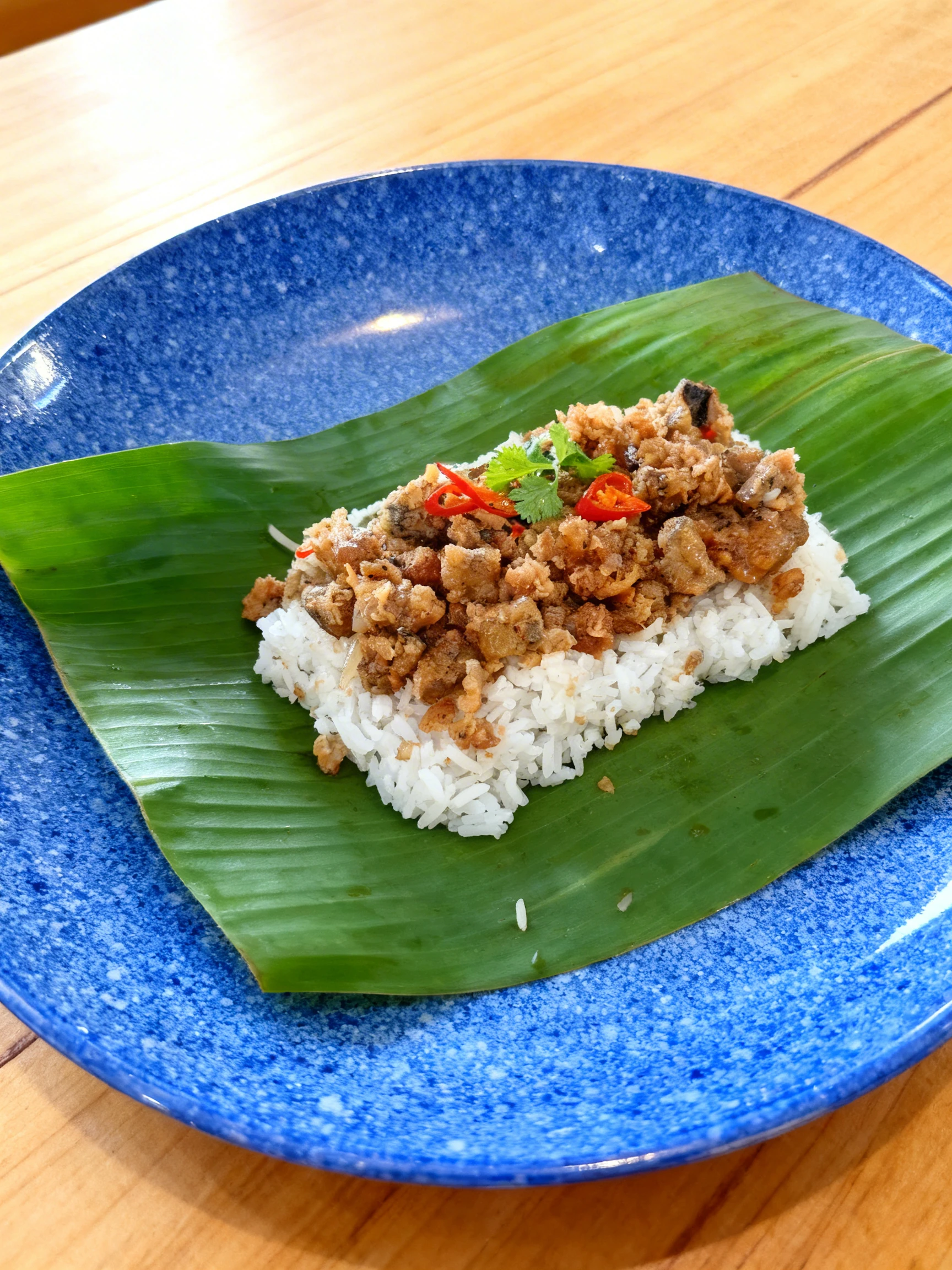 Crispy fried fish pieces or pork nuggets served over steamed white rice on a banana leaf, garnished with sliced red chilies and fresh cilantro.