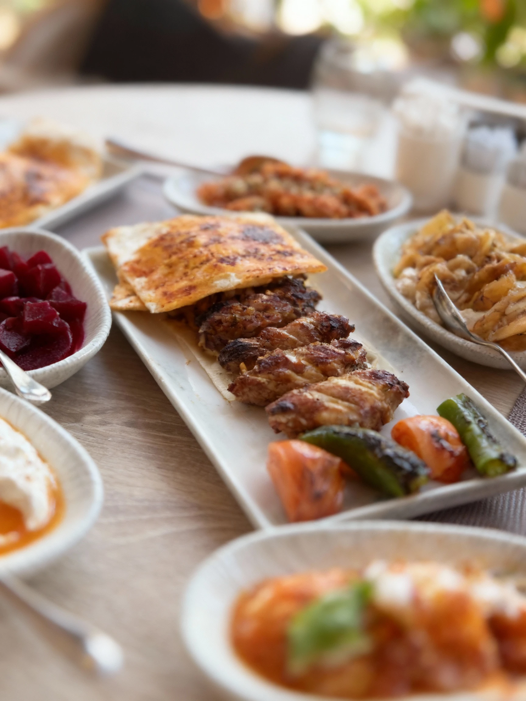 Turkish dinner spread featuring Adana kebab and grilled chicken wings served with flatbread, grilled peppers, and tomatoes, accompanied by various mezze includi