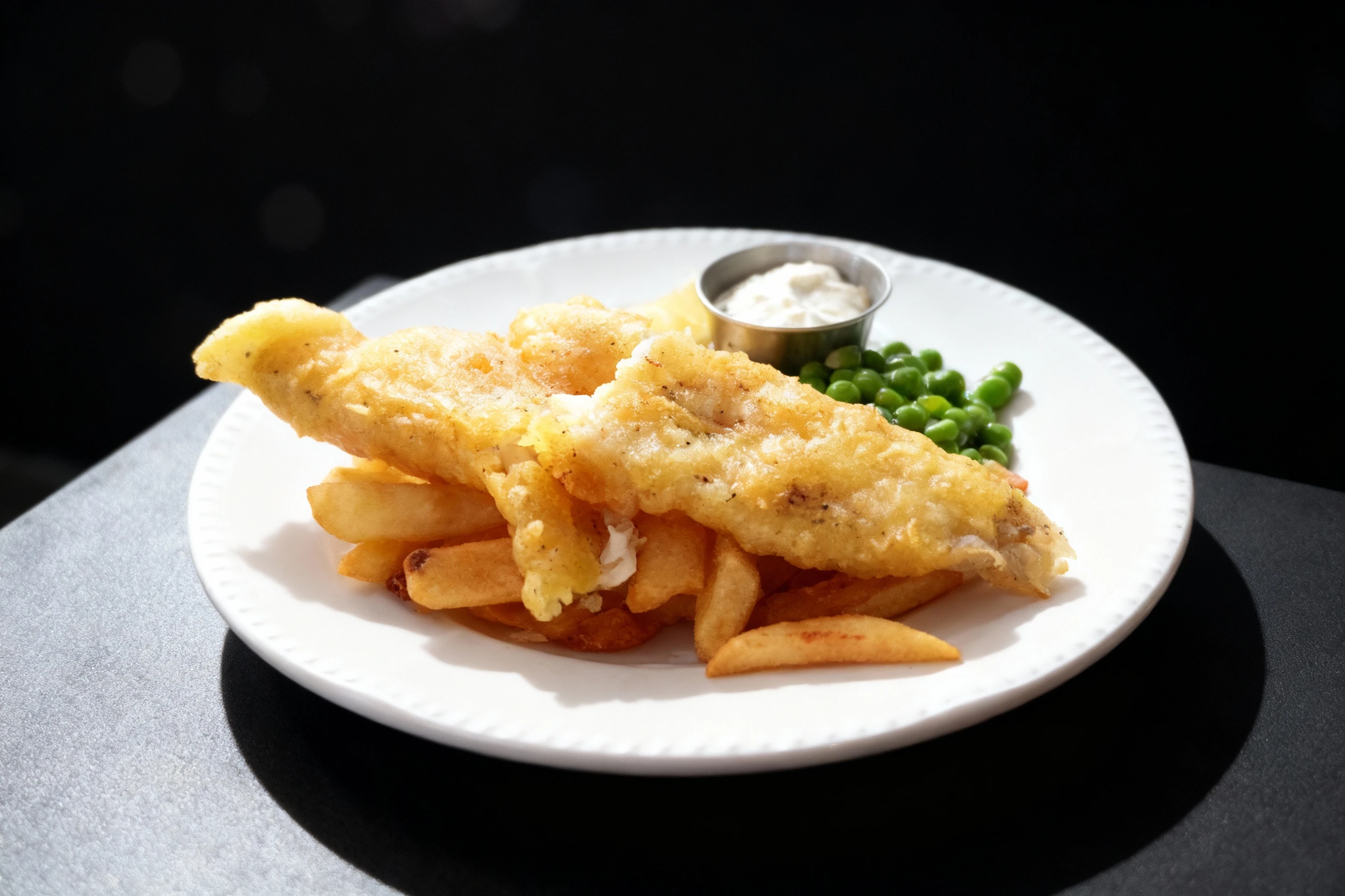 Battered fish and chips served with a side of green peas, a lemon wedge, and a small metal ramekin of tartar sauce.