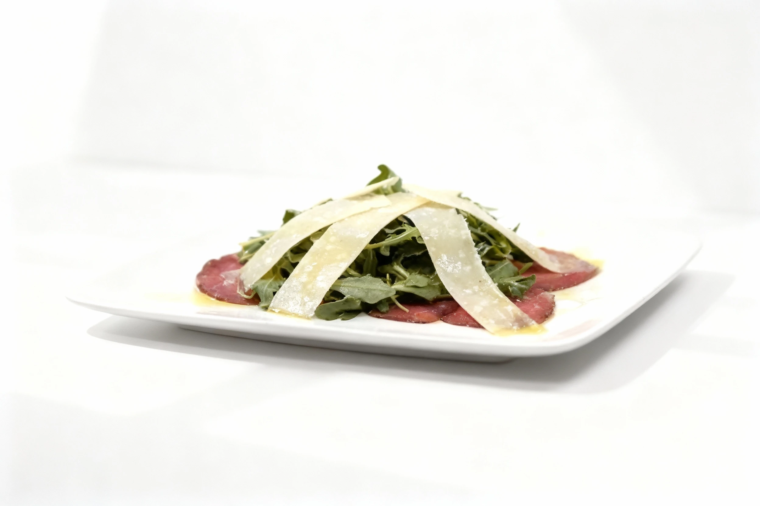Beef carpaccio consisting of thin slices of raw beef topped with fresh arugula leaves and large shavings of parmesan cheese, finished with an oil dressing.