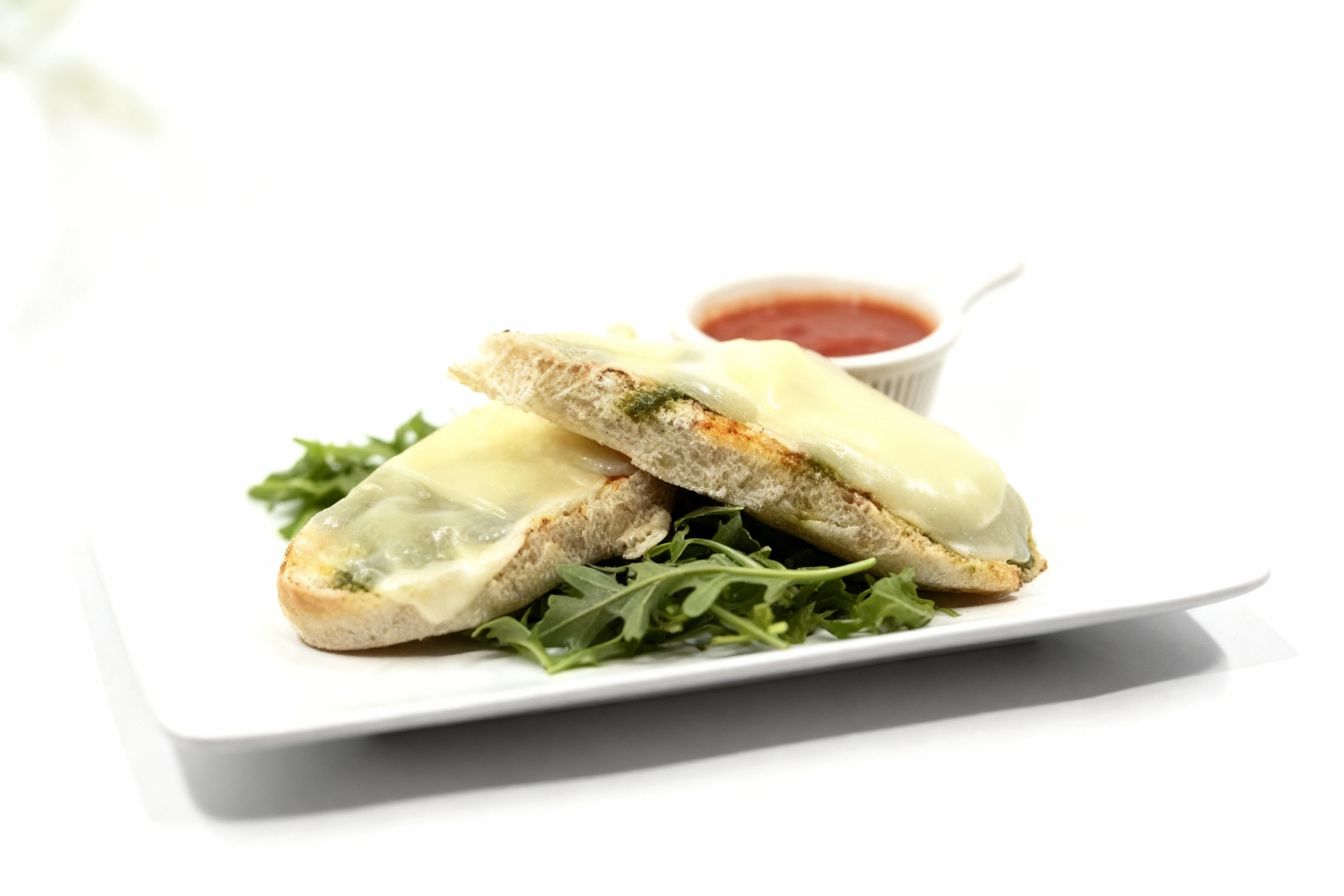 Two slices of toasted bread topped with melted mozzarella cheese and pesto, served on a bed of fresh arugula with a side of tomato dipping sauce.