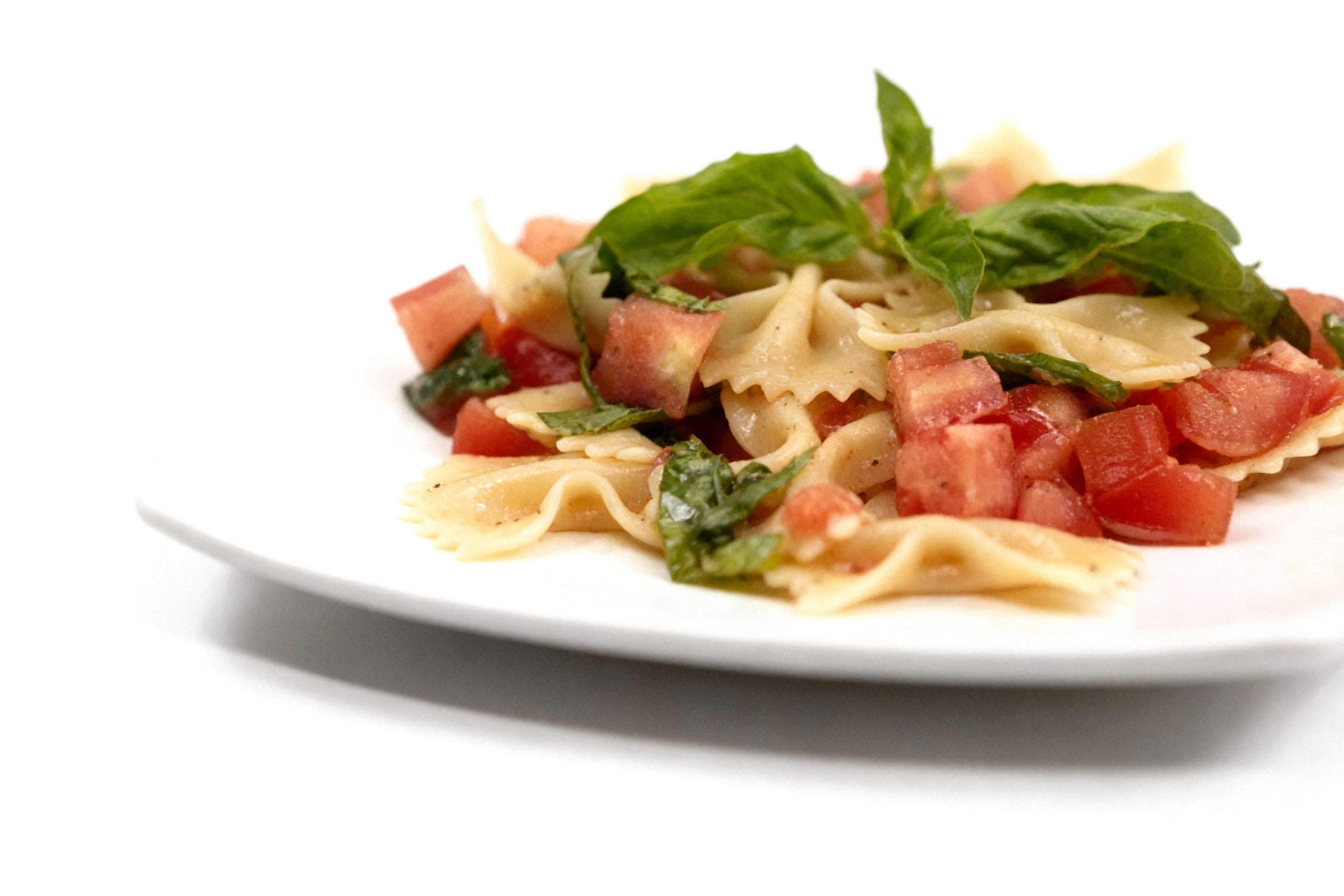 Farfalle pasta salad mixed with diced tomatoes and fresh basil leaves on a white square plate.