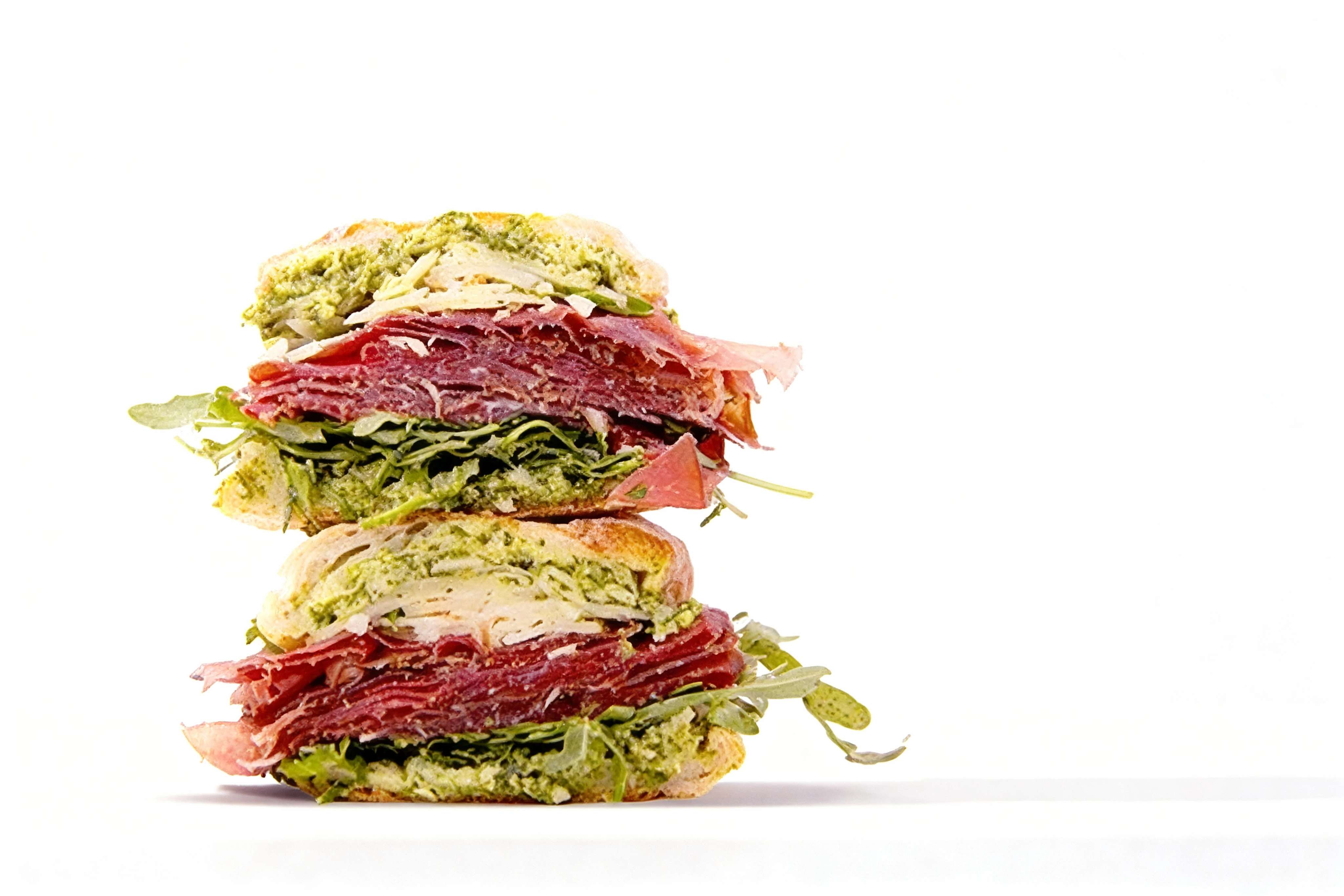 A stacked sandwich on crusty bread featuring thin slices of cured meat, fresh arugula, pesto spread, and shaved parmesan cheese.