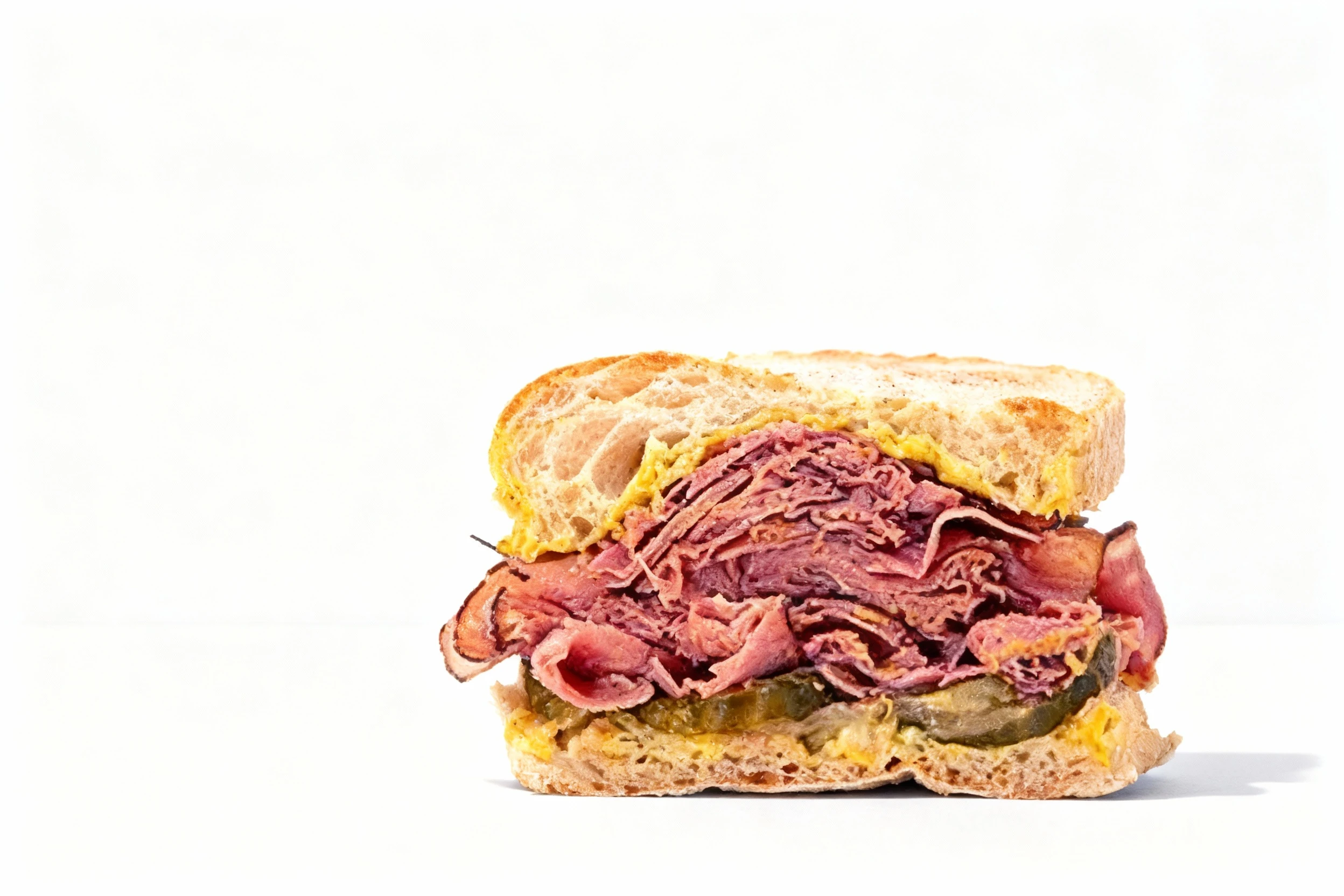A stacked pastrami sandwich cut in half on toasted sourdough bread with sliced pickles and yellow mustard.