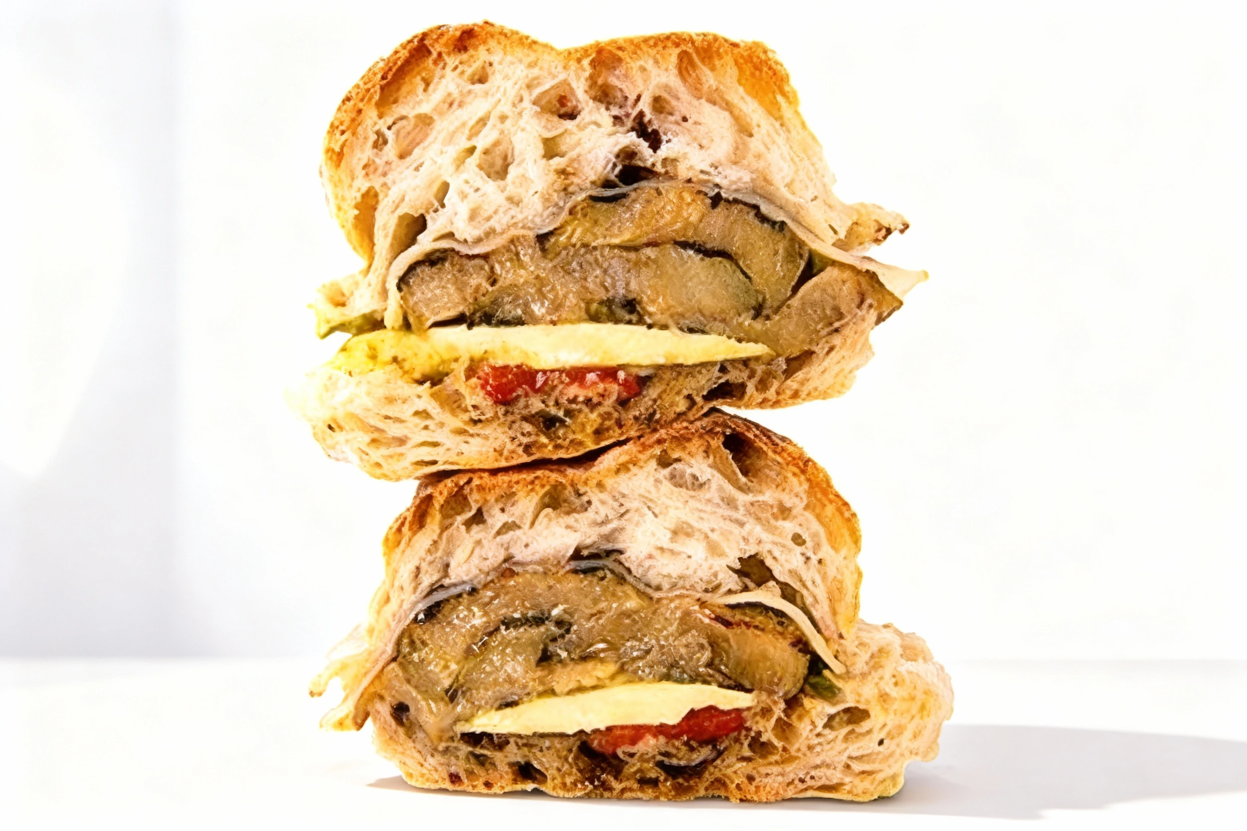 A toasted sandwich made with crusty bread, containing layered fillings of roasted eggplant, melted cheese, and sliced red tomato.
