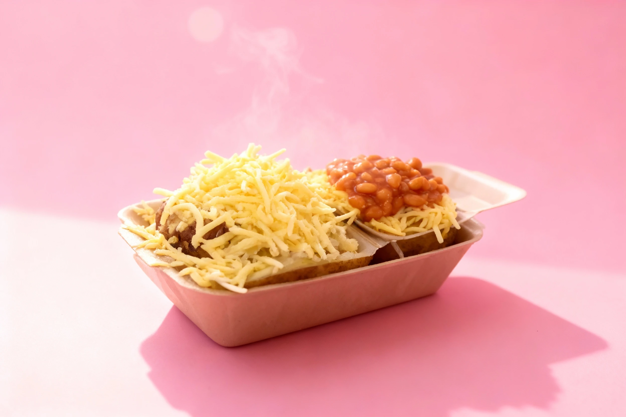 A jacket potato served in a takeaway container, topped with a large portion of shredded cheddar cheese and steaming baked beans.