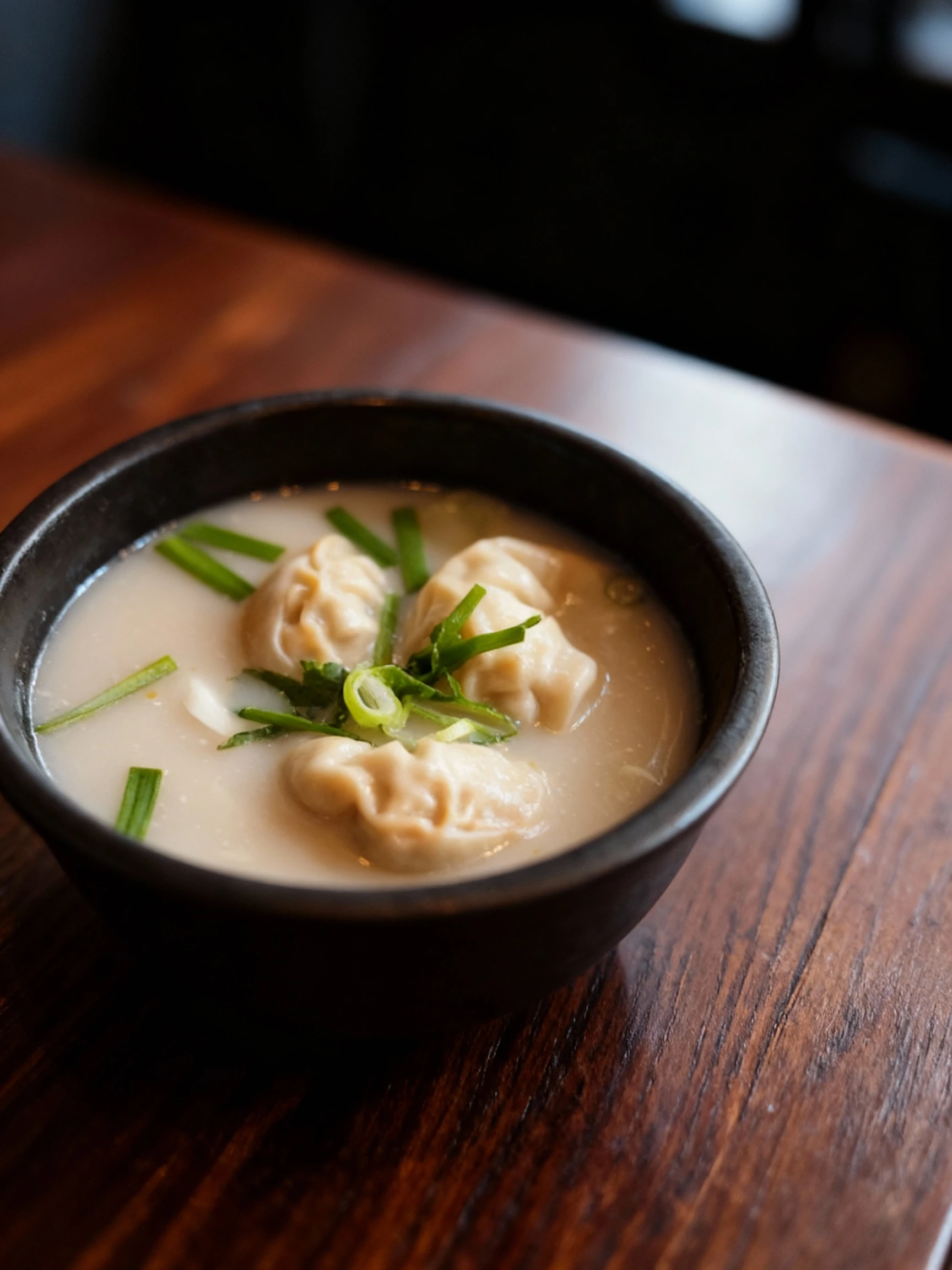 A bowl of dumpling soup featuring steamed dumplings in a milky white bone broth, garnished with sliced scallions and garlic chives.