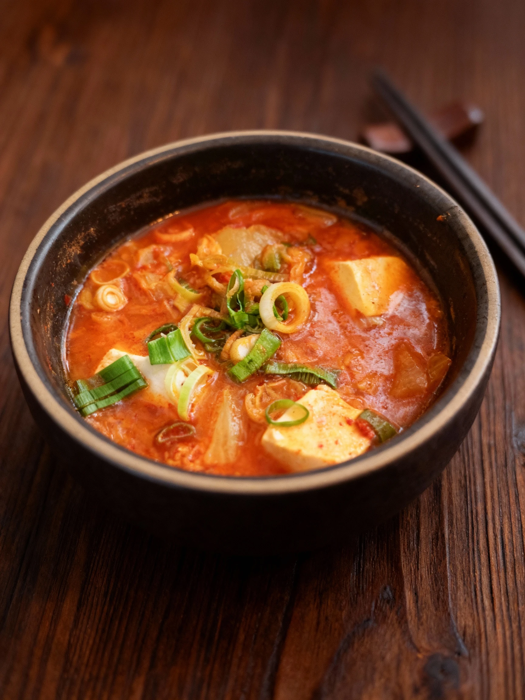 A bowl of Korean Kimchi-jjigae featuring cubes of soft tofu and fermented cabbage in a spicy broth, topped with sliced green onions.