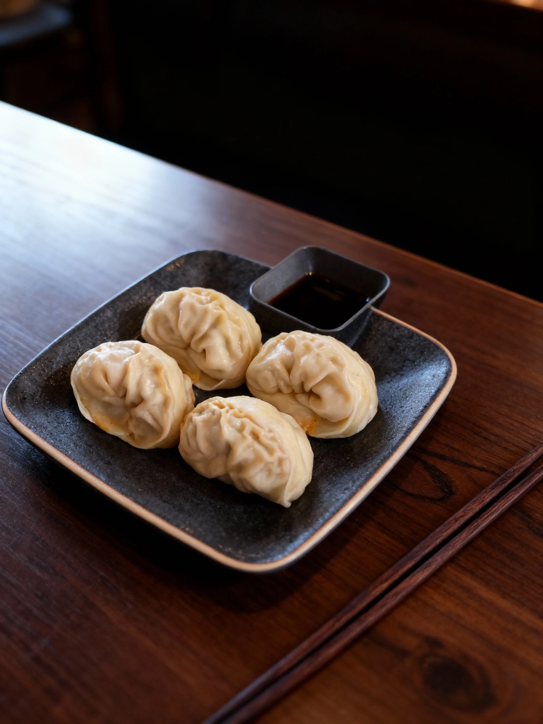 Four steamed dumplings with pleated dough wrappers arranged on a grey square plate with a built-in sauce compartment.