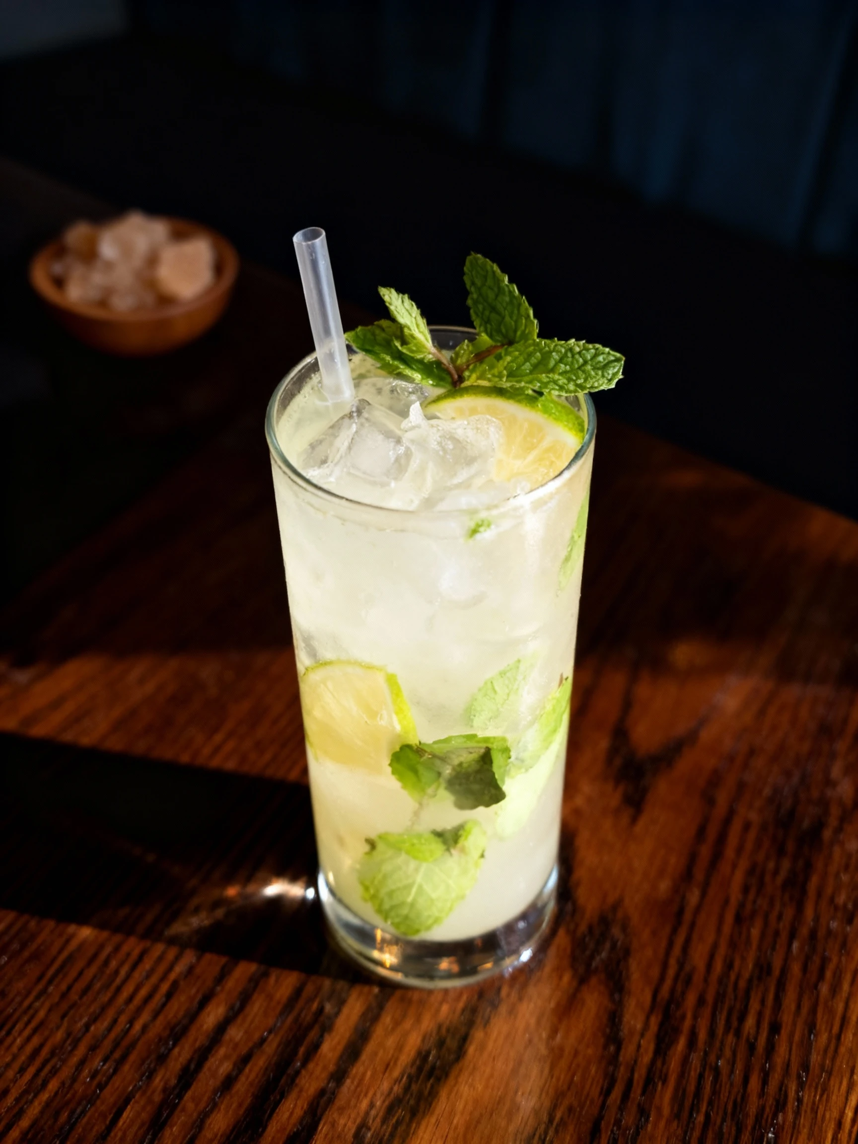 A mojito cocktail served in a highball glass with muddled mint leaves, lime juice, and a plastic straw.