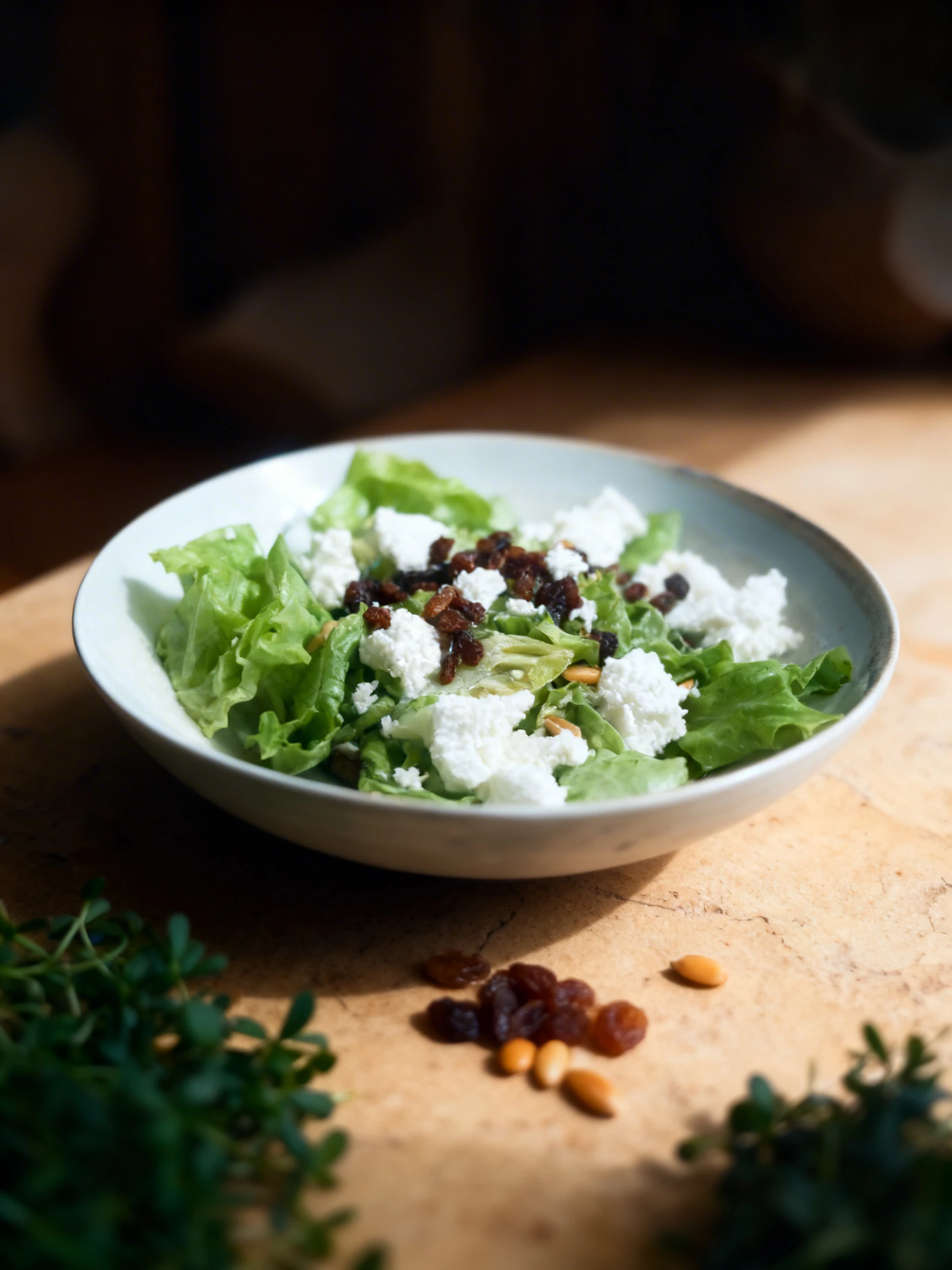A green salad featuring butter lettuce leaves topped with crumbles of goat cheese, raisins, and pine nuts in a light-colored ceramic bowl.