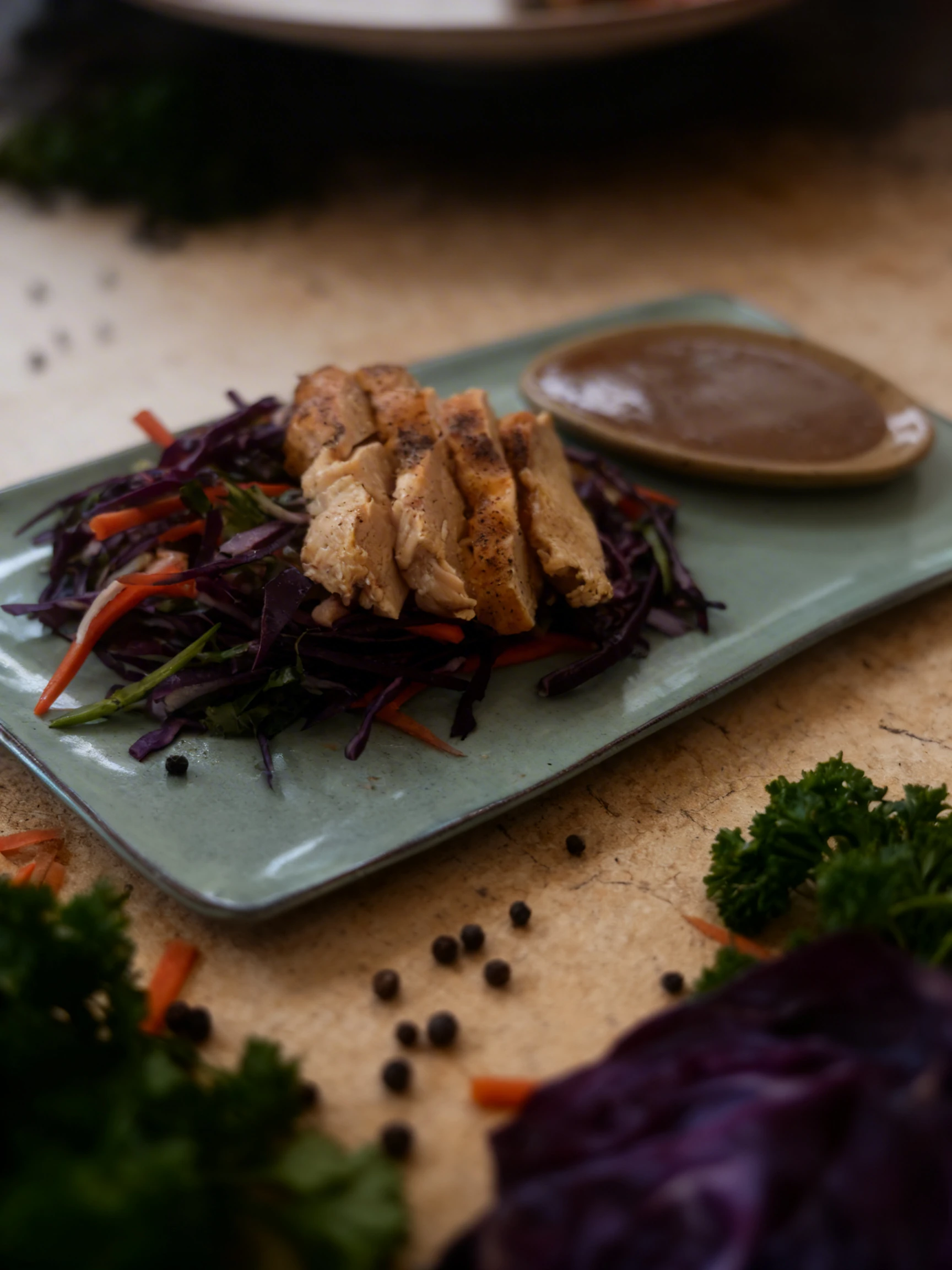Slices of roasted chicken breast seasoned with black pepper, served on a bed of shredded red cabbage and carrot slaw with a side of brown dipping sauce.