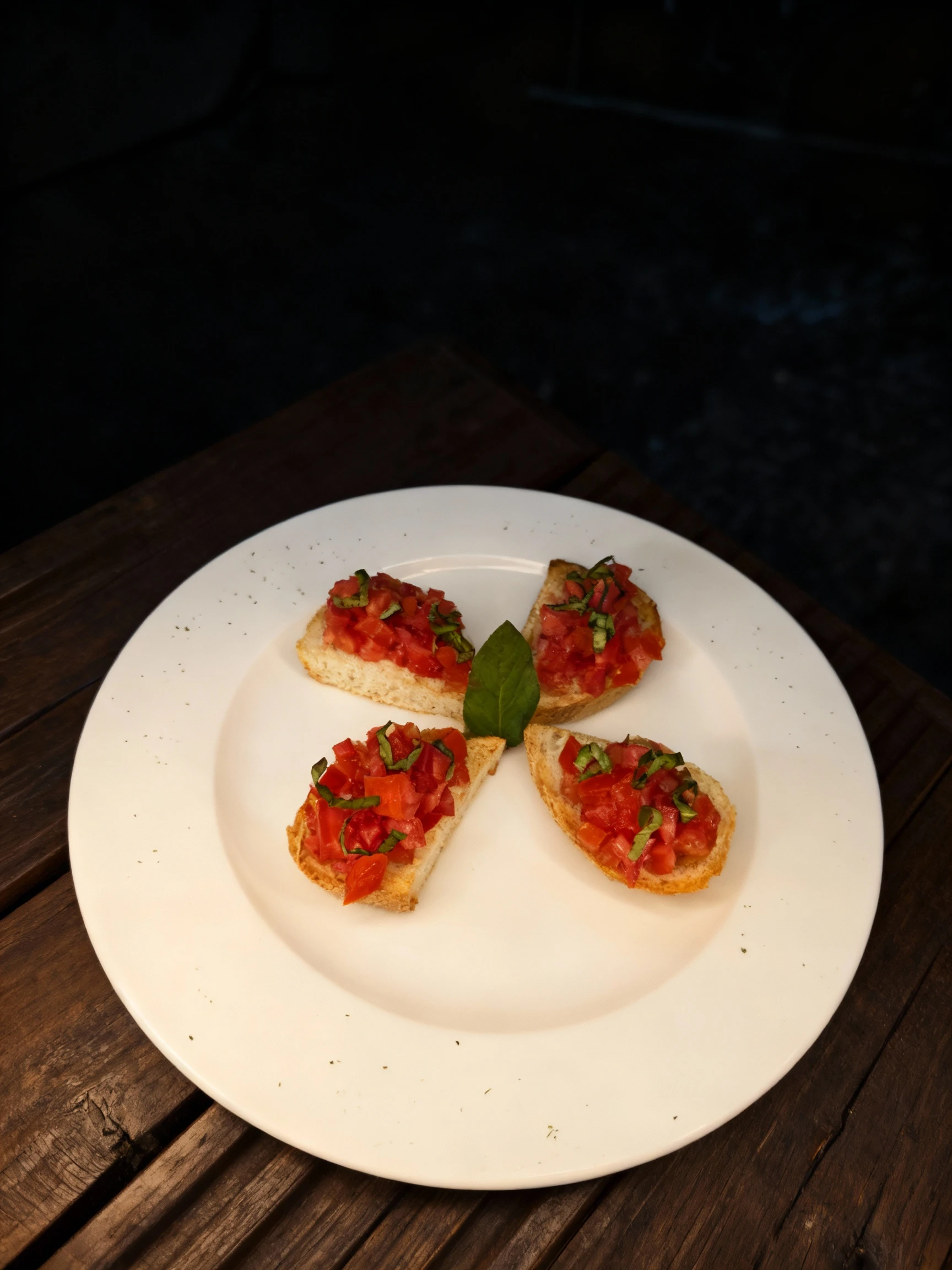 Four pieces of tomato bruschetta topped with diced tomatoes and thinly sliced fresh basil, served on a white plate with a central basil leaf garnish.