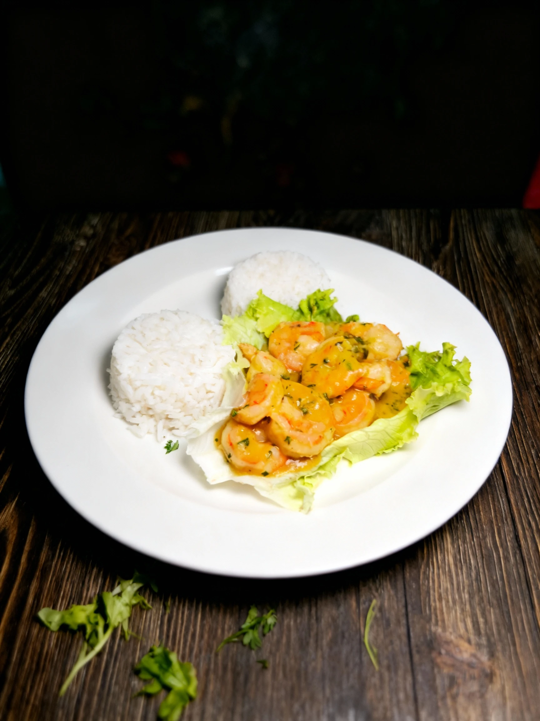 A dish featuring peeled shrimp coated in a vibrant yellow, herbed sauce, served alongside two molded portions of plain white rice and garnished with fresh lettu