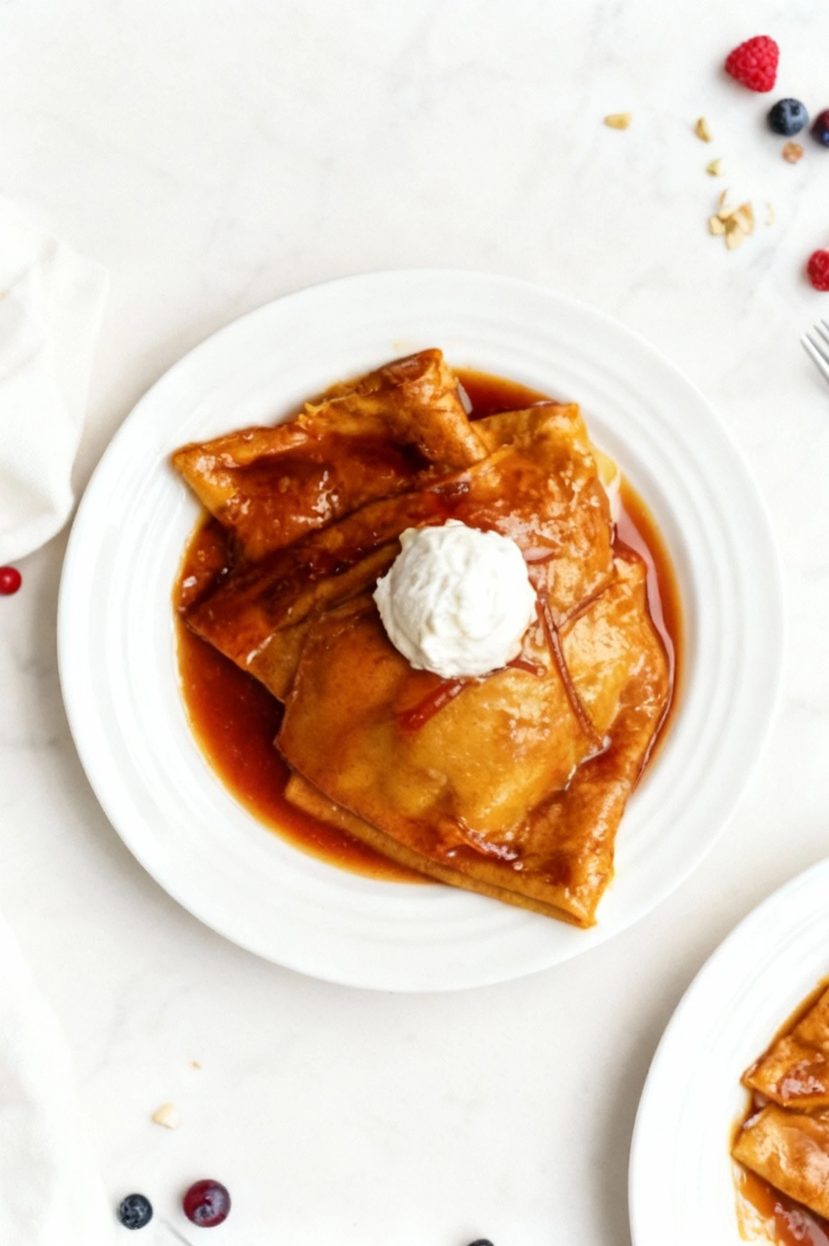 Folded crepes or thin pancakes are served on a white plate, drenched in a glossy, rich caramel or reddish-brown syrup, and topped with a large dollop of whipped