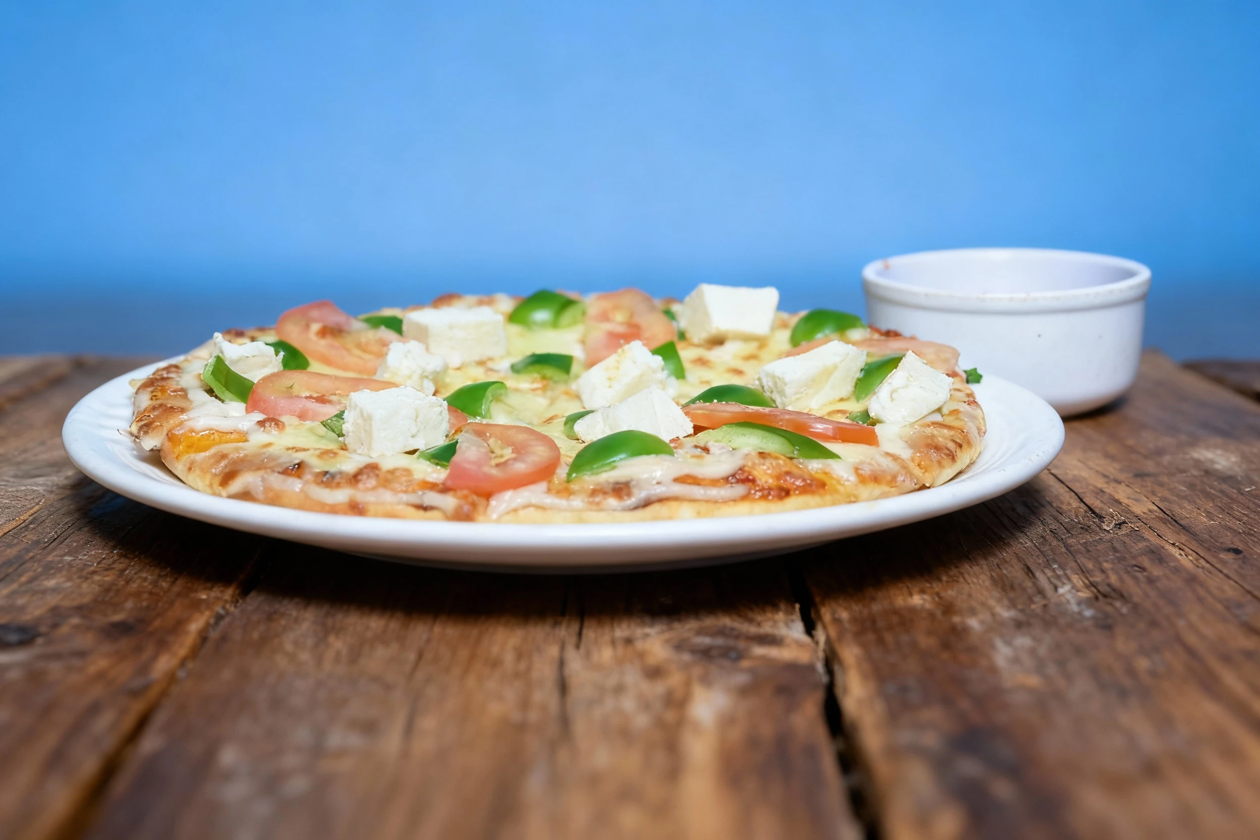 This is a vegetarian pizza topped with melted mozzarella cheese, large chunks of paneer or feta, green bell peppers, and sliced tomatoes, presented on a white p