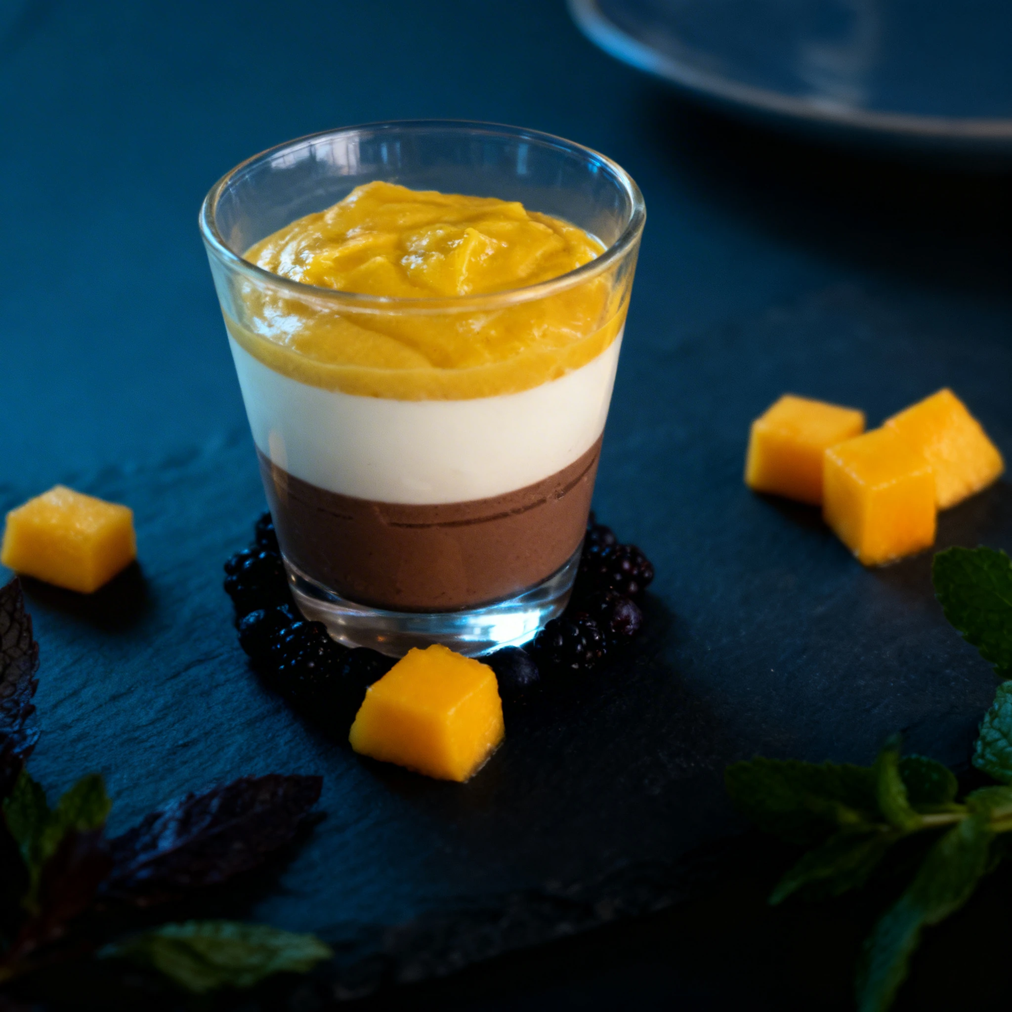 Layered dessert in a glass featuring chocolate mousse at the base, a middle layer of white cream or panna cotta, and topped with a mango or fruit puree.
