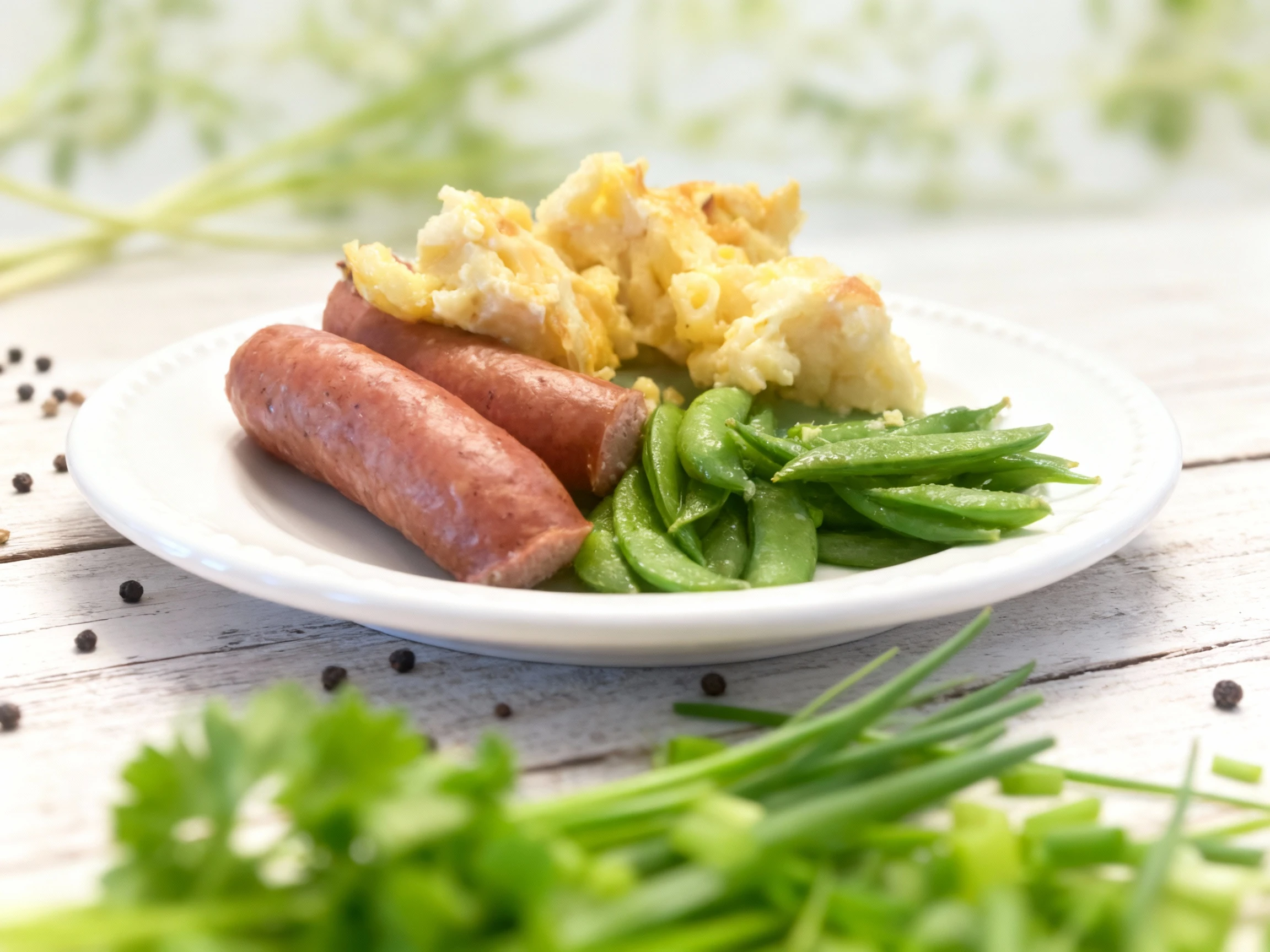 A simple meal featuring two whole smoked sausages, a scoop of baked macaroni and cheese with a golden top, and a side of whole sugar snap peas.