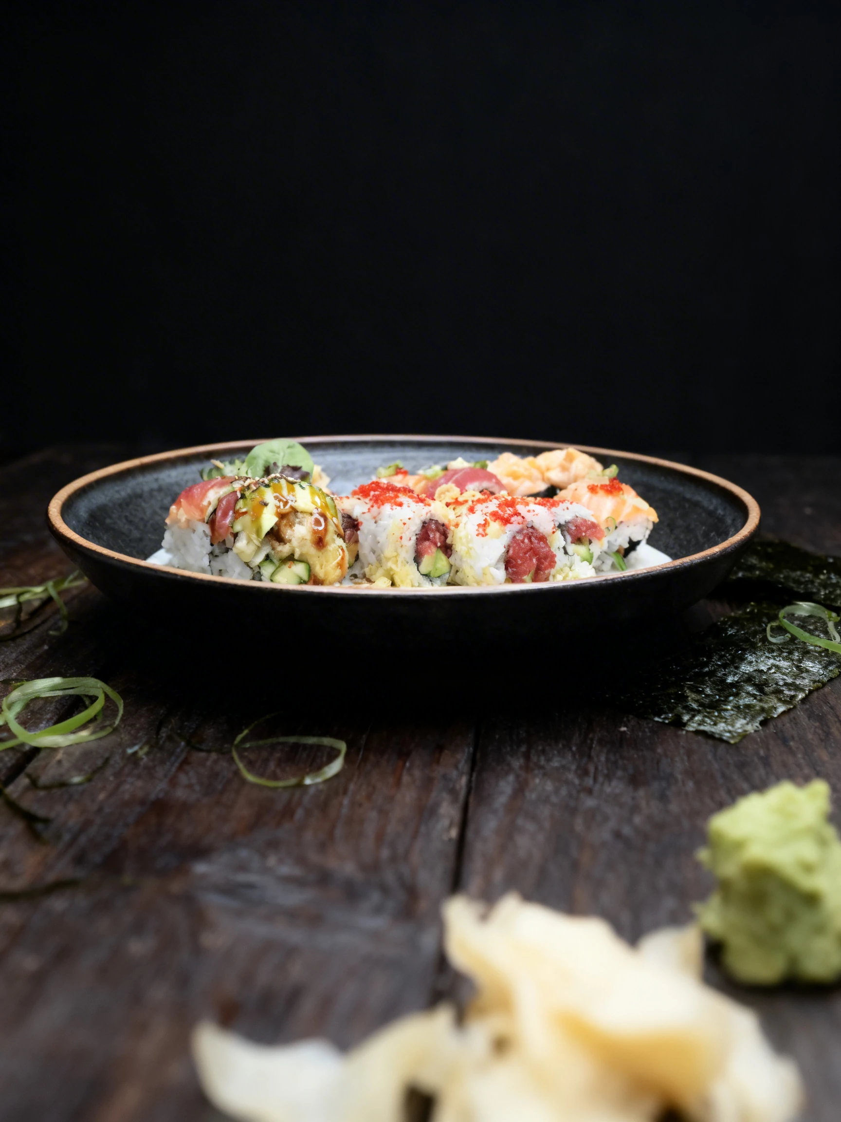 A platter of various sushi rolls featuring tuna, salmon, cucumber, and avocado, topped with red tobiko and green onions, with one piece held by chopsticks.