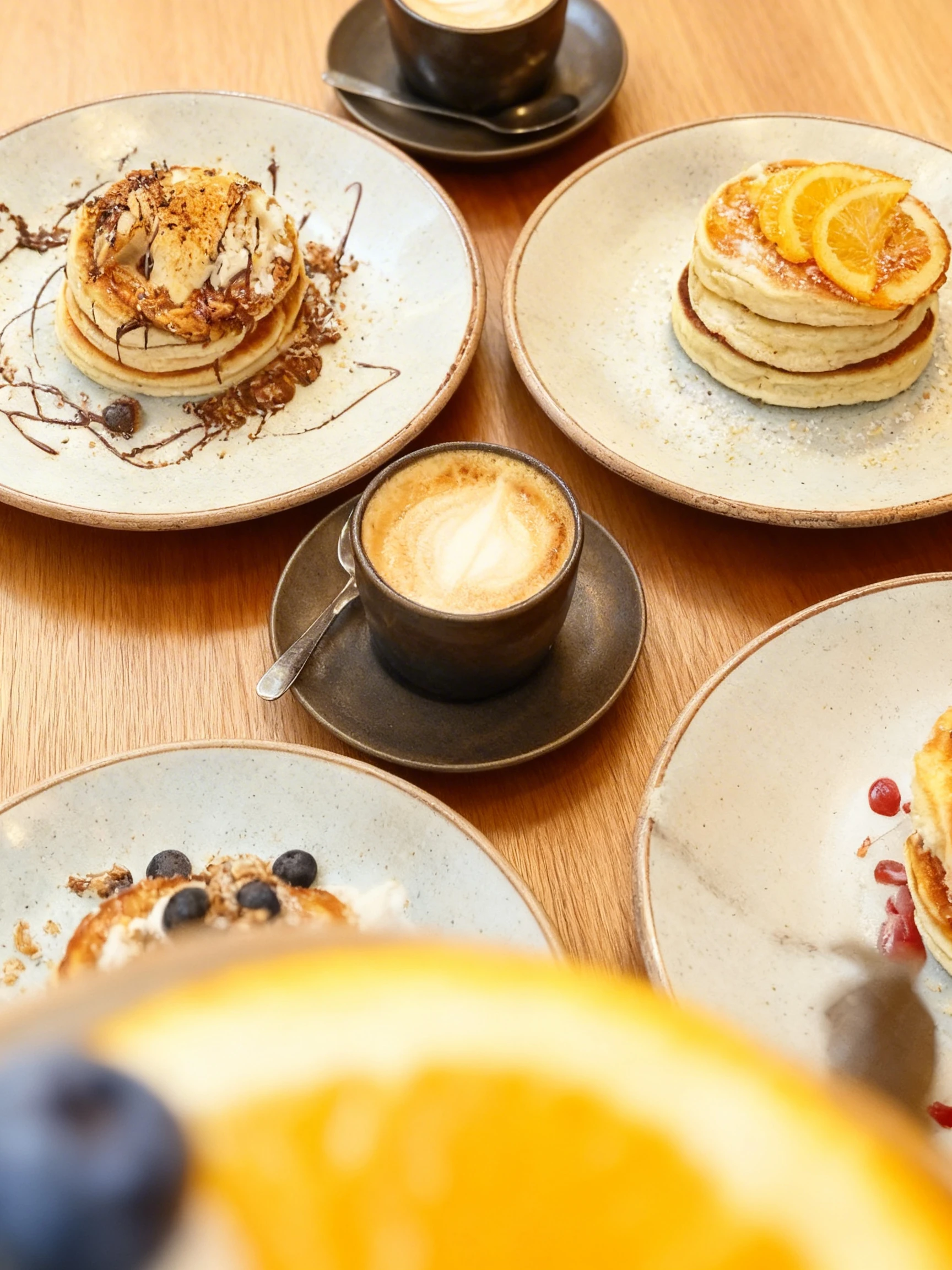 A breakfast spread featuring four stacks of pancakes with various toppings including sliced bananas with chocolate drizzle, fresh blueberries with granola, stra