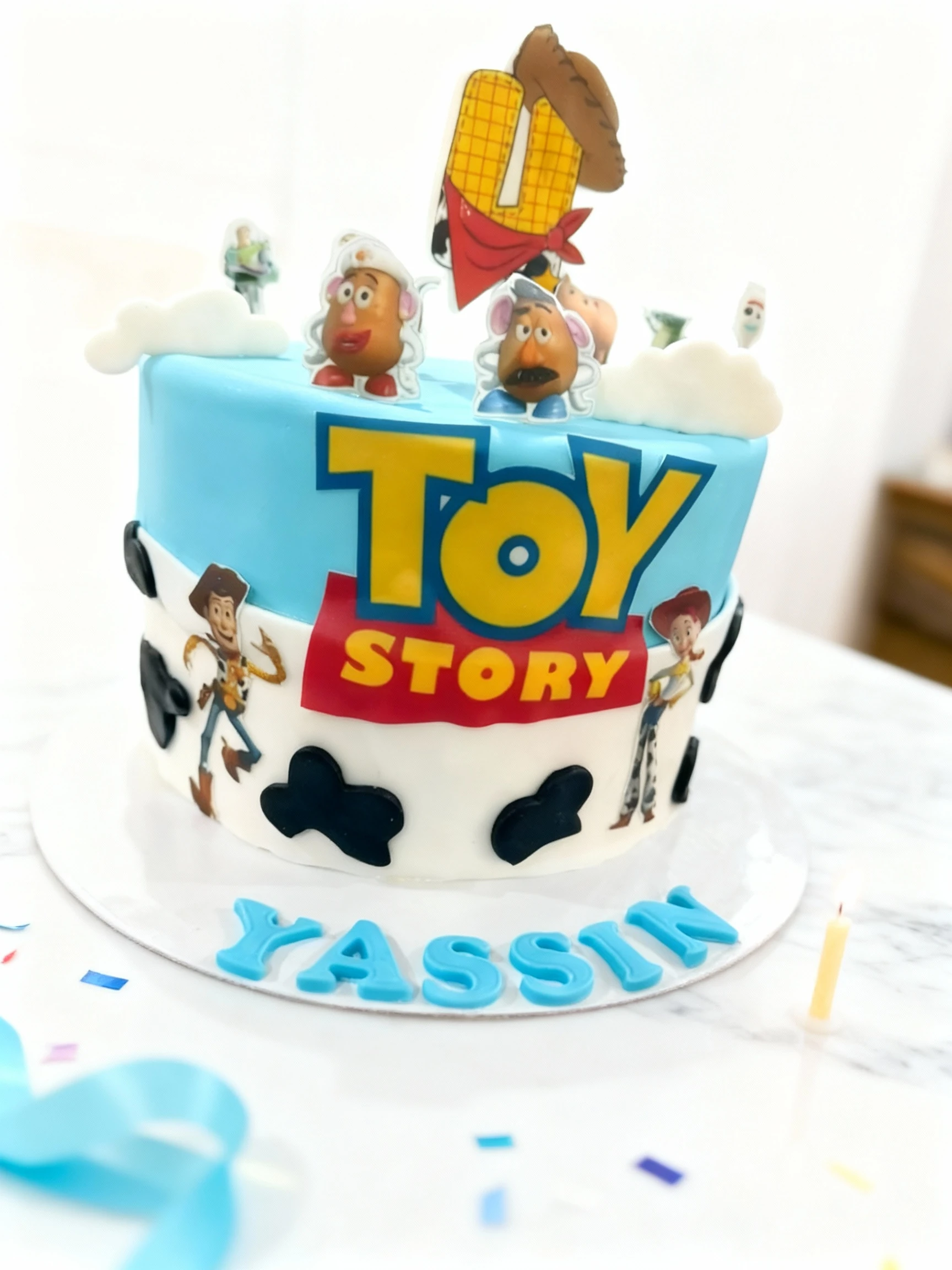 A two-tiered birthday cake featuring a light blue top layer displaying the "Toy Story" logo and character cutouts, and a white bottom layer decorated with cow-p