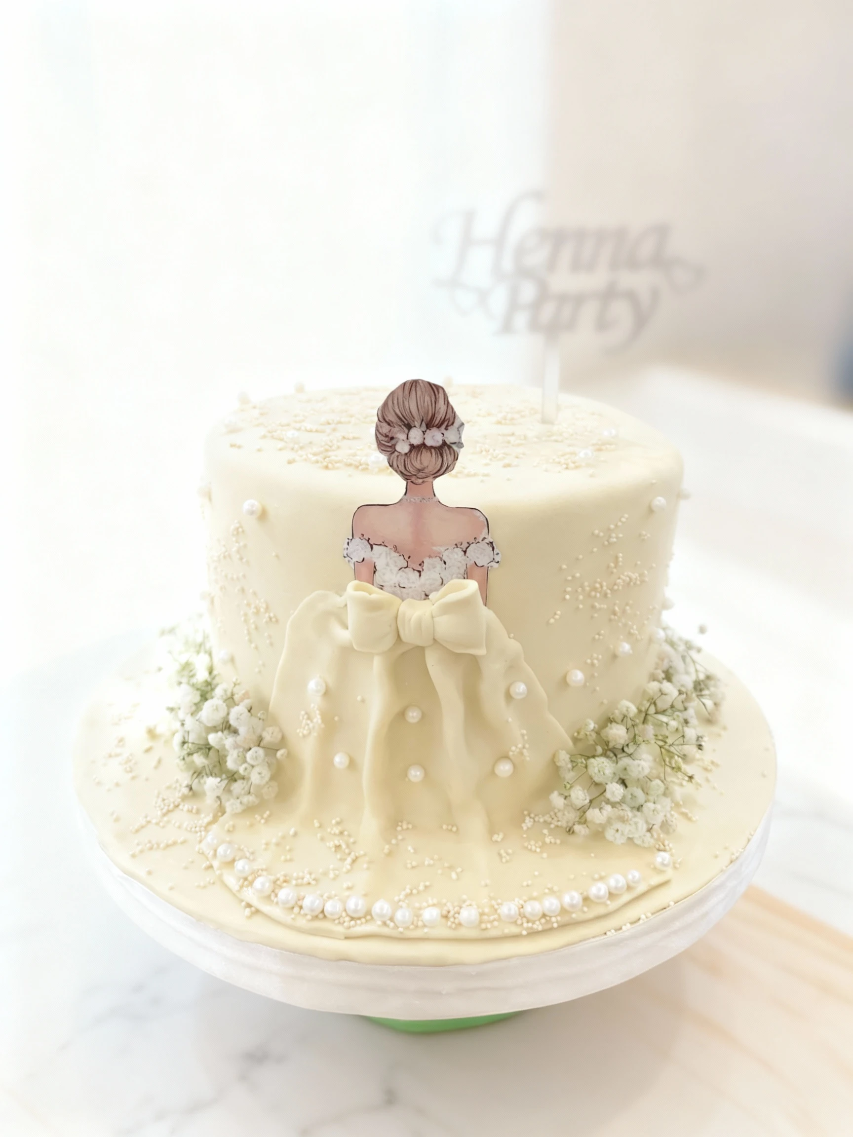 A cream-colored, multi-layered celebration cake decorated with edible pearls, sprinkles, and clusters of baby's breath flowers. The cake features a printed cuto