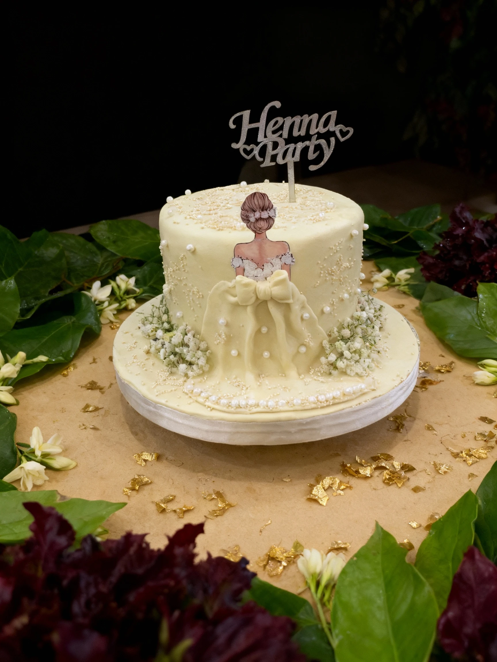 A celebratory two-tiered cake covered in cream-colored fondant, elaborately decorated with edible white pearls, bead sprinkles, and small white flowers, featuri