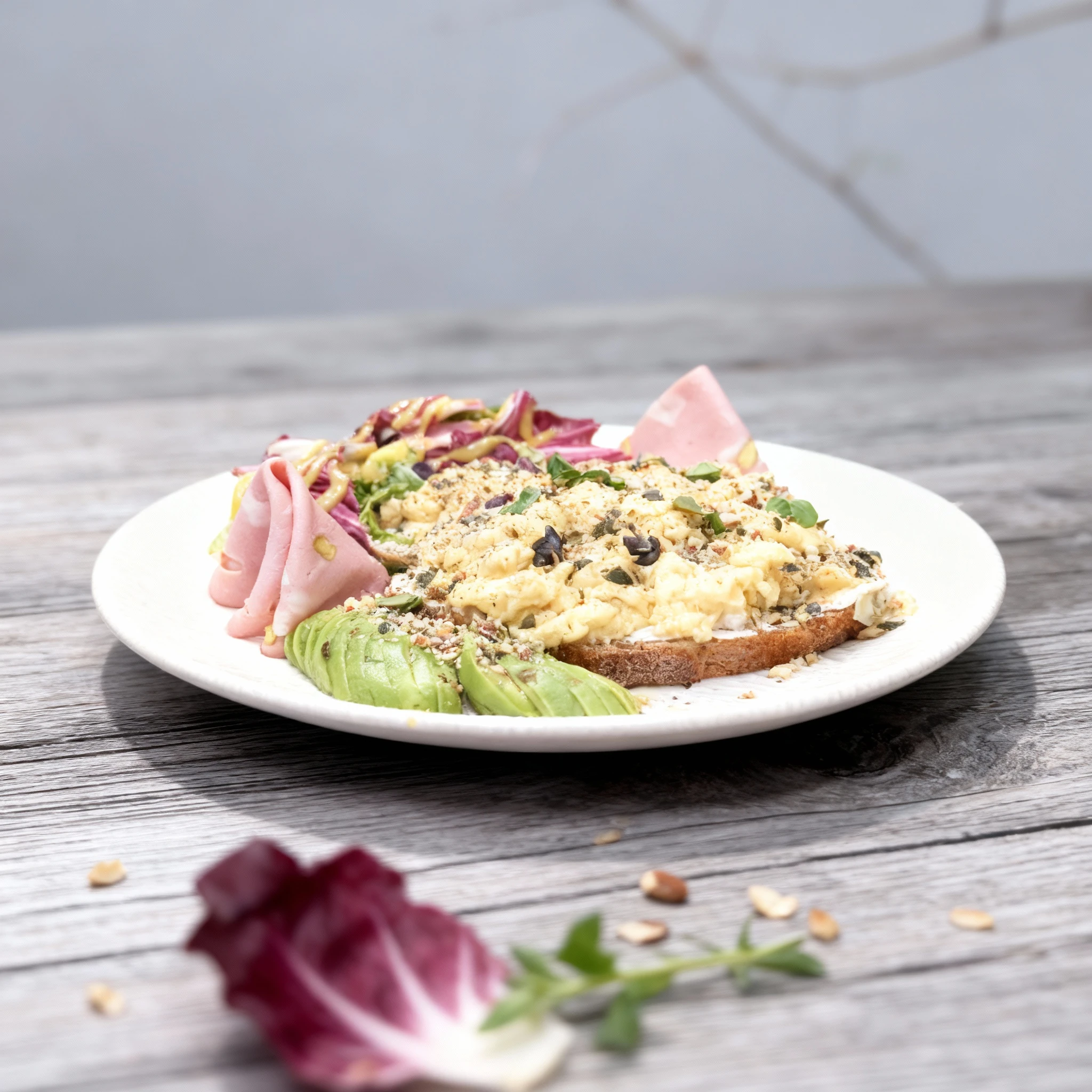 Breakfast toast topped with creamy scrambled eggs, crushed nuts, and seeds, served with sliced avocado, mortadella, and a radicchio salad drizzled with mustard