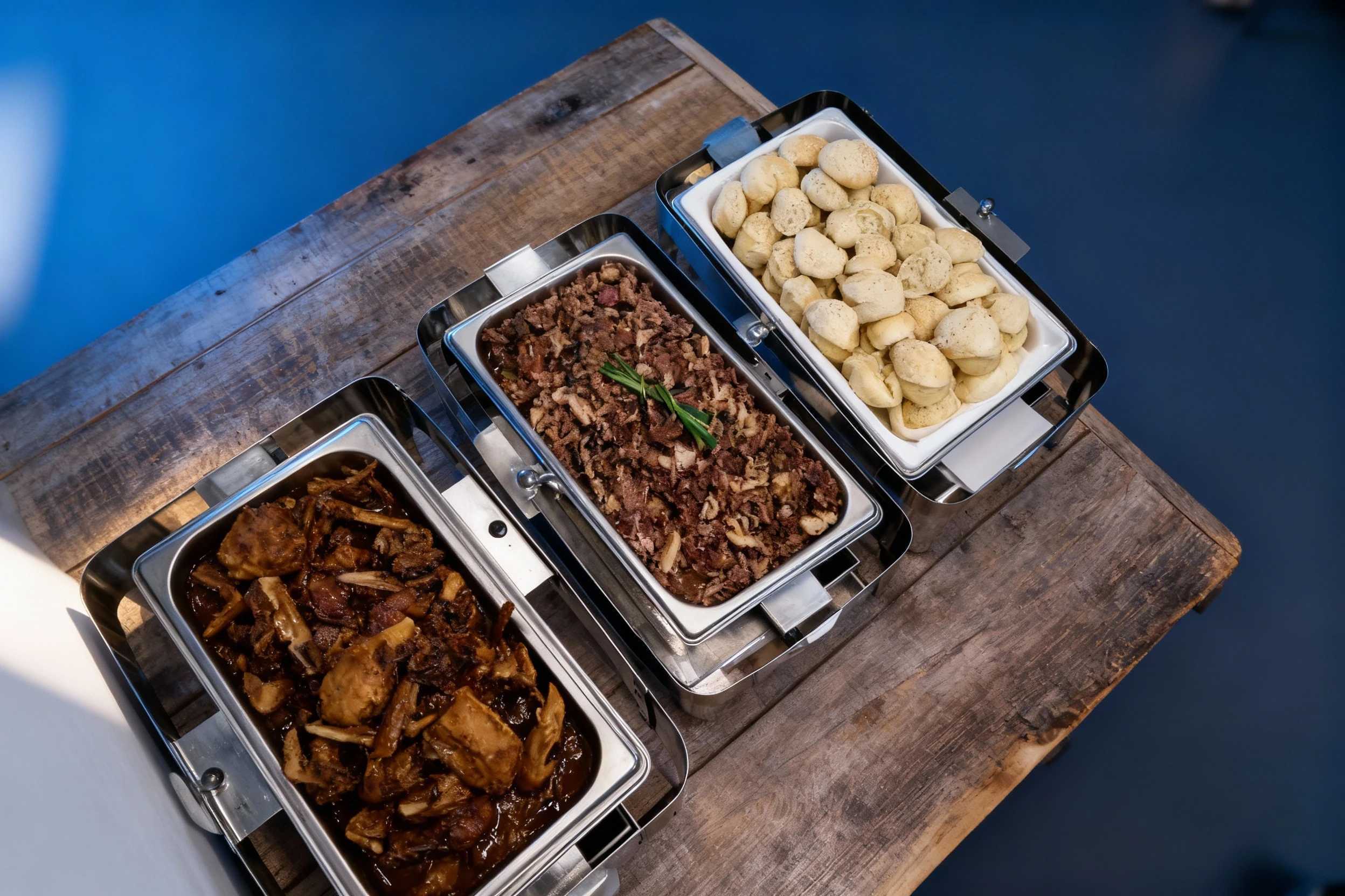 This is a buffet setup featuring several stainless steel chafing dishes containing various foods, including a foreground dish of sauced meat pieces, a central t