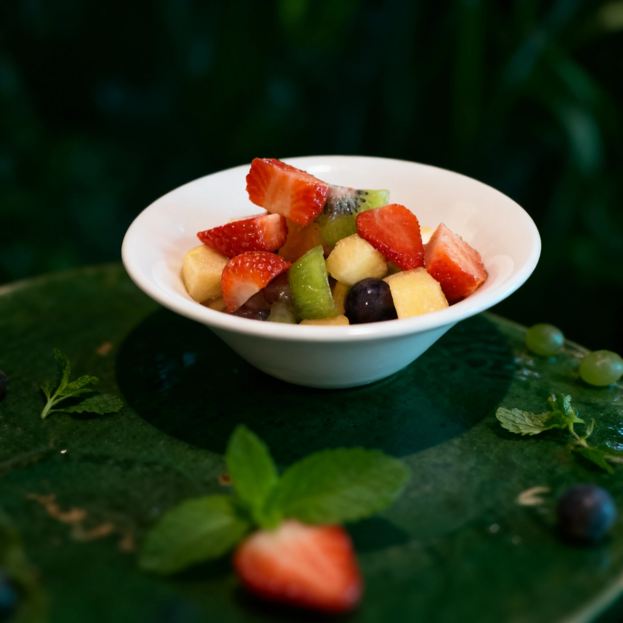 A fresh fruit salad containing sliced strawberries, kiwi, melon, and grapes served in a white bowl atop a red paper doily.
