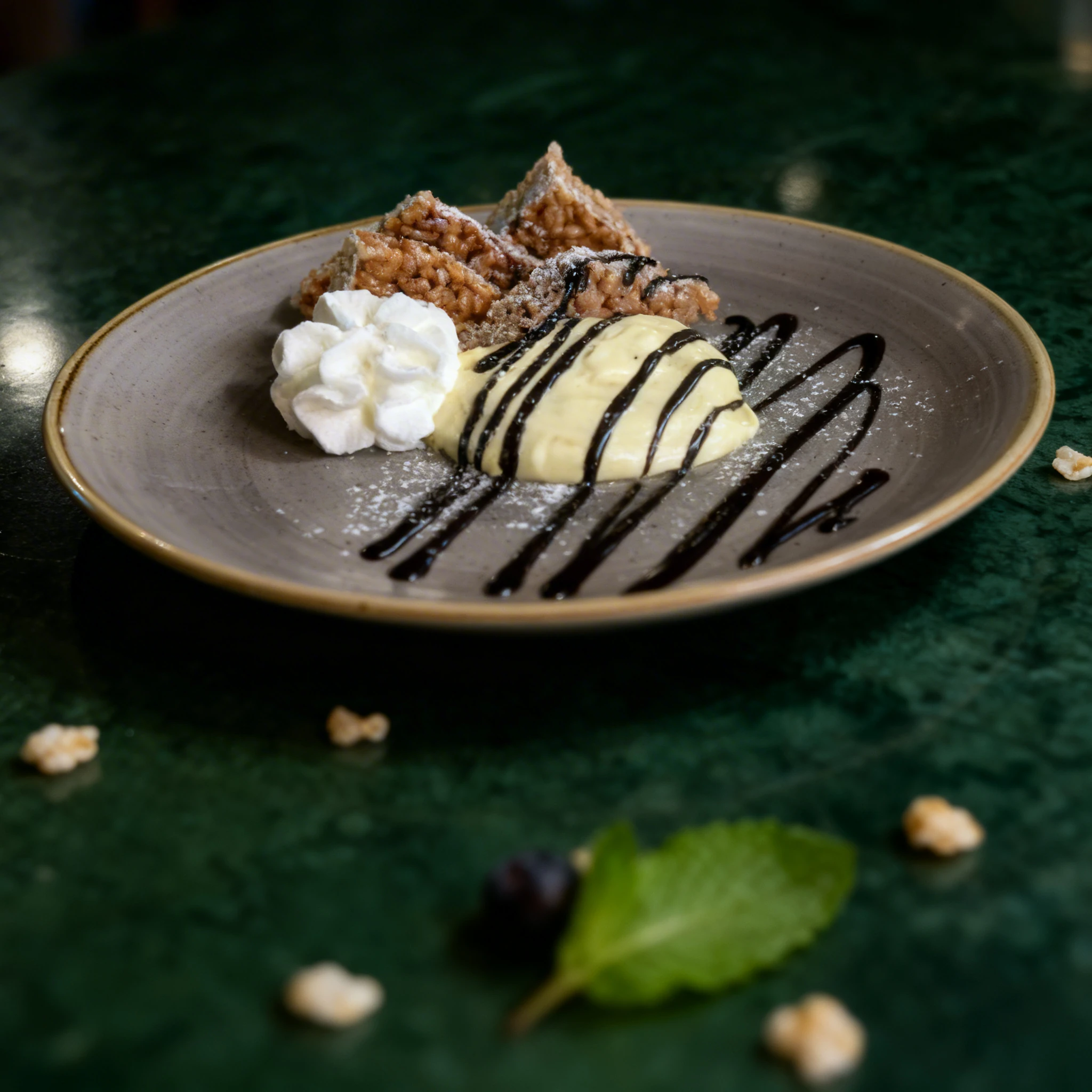 A dessert plate featuring puffed rice treats, a dollop of whipped cream, and a scoop of vanilla custard or ice cream drizzled with chocolate sauce and a dusting
