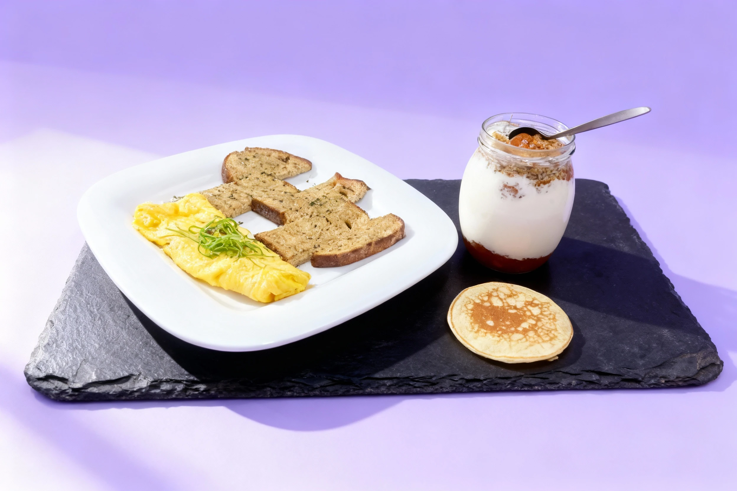 A breakfast spread featuring a folded omelet garnished with sliced green herbs, several slices of herbed toast, a single pancake, and a layered yogurt parfait s