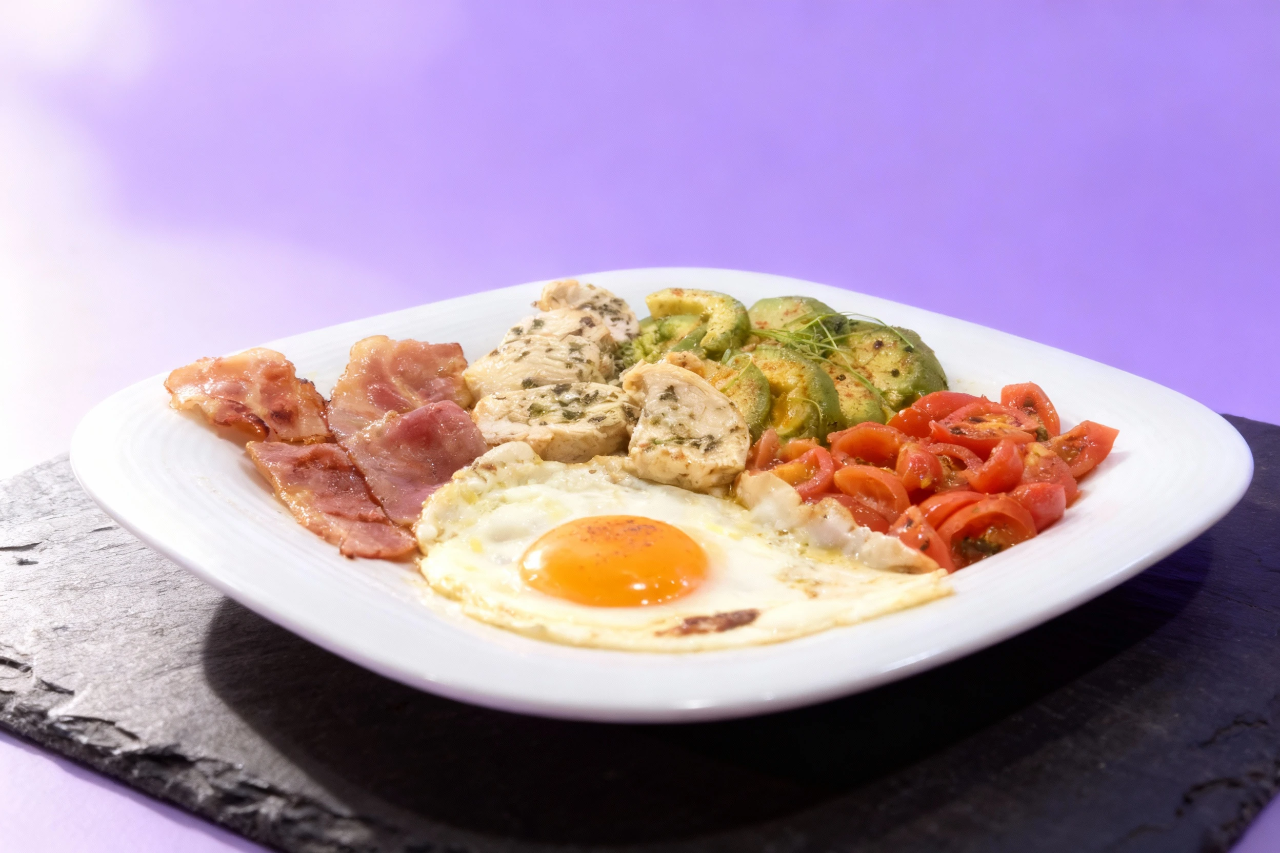 A protein plate featuring a sunny-side-up egg, strips of bacon, herbed chicken breast pieces, seasoned sliced avocado, and sautéed halved cherry tomatoes.