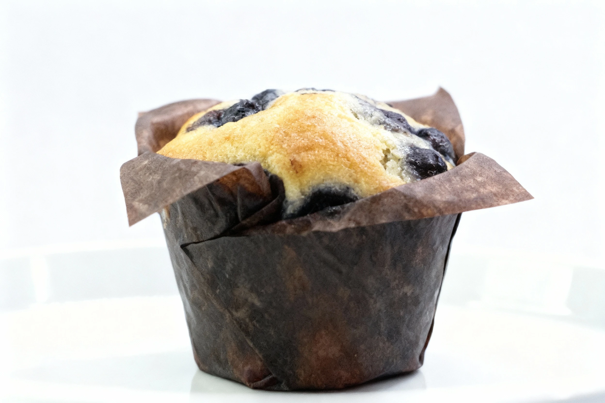 A golden-baked blueberry muffin topped with visible berries, presented in its tall brown paper liner and centered on a plain white serving plate.
