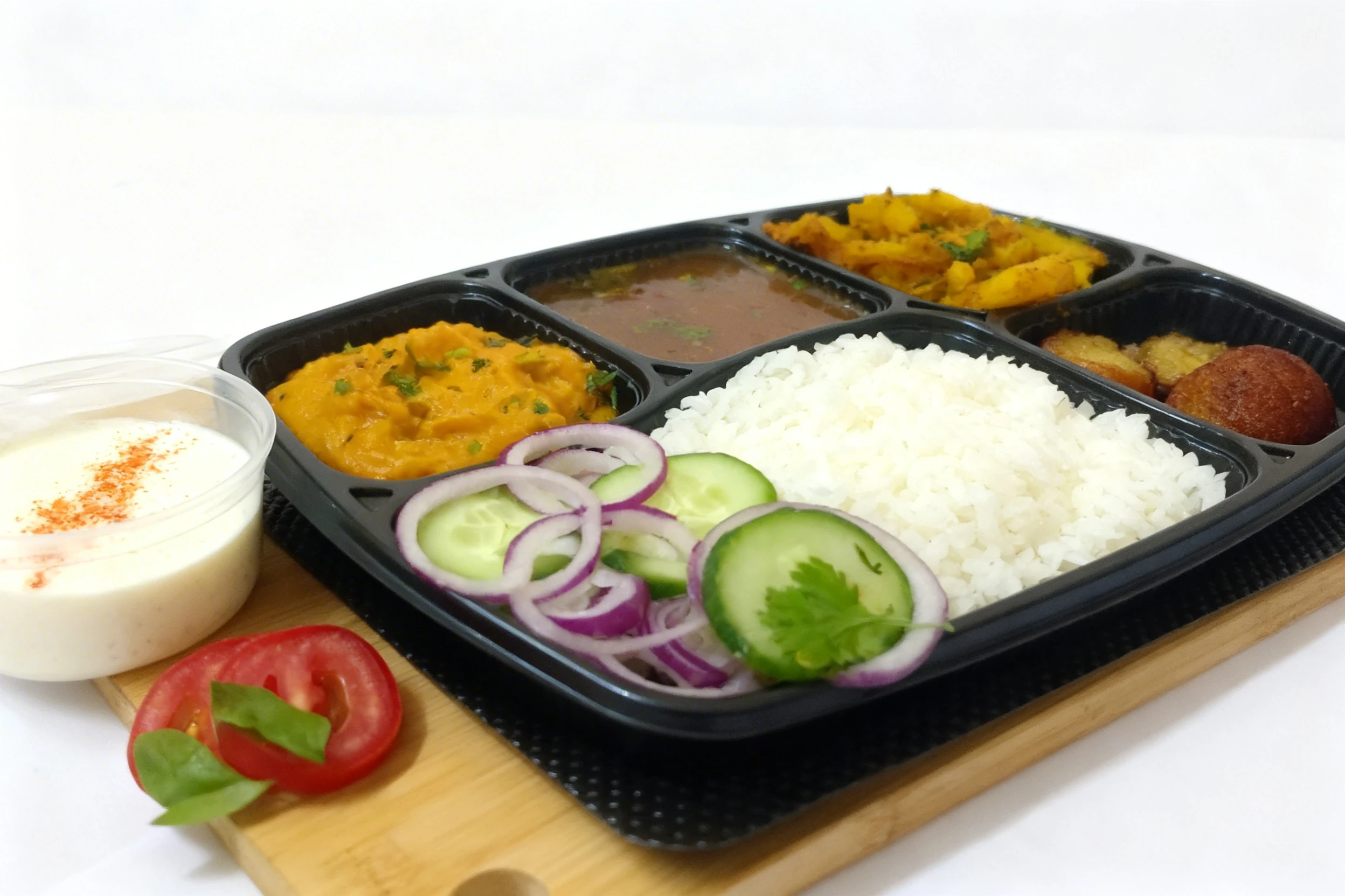 An Indian thali meal featuring spiced potatoes, dal, a thick yellow curry, steamed white rice, gulab jamun, and a side of raita. Fresh salad garnishes including