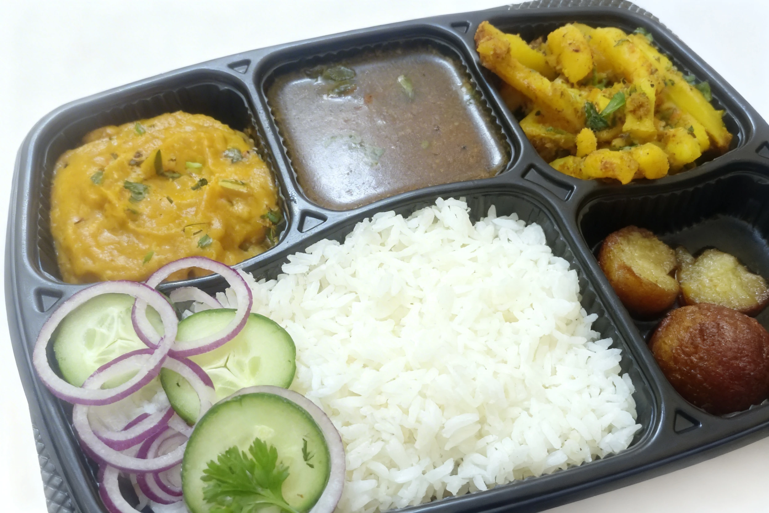 Indian thali meal in a black bento tray featuring steamed white rice, yellow dal, vegetable curry, spiced potatoes, and gulab jamun with a side of cucumber and